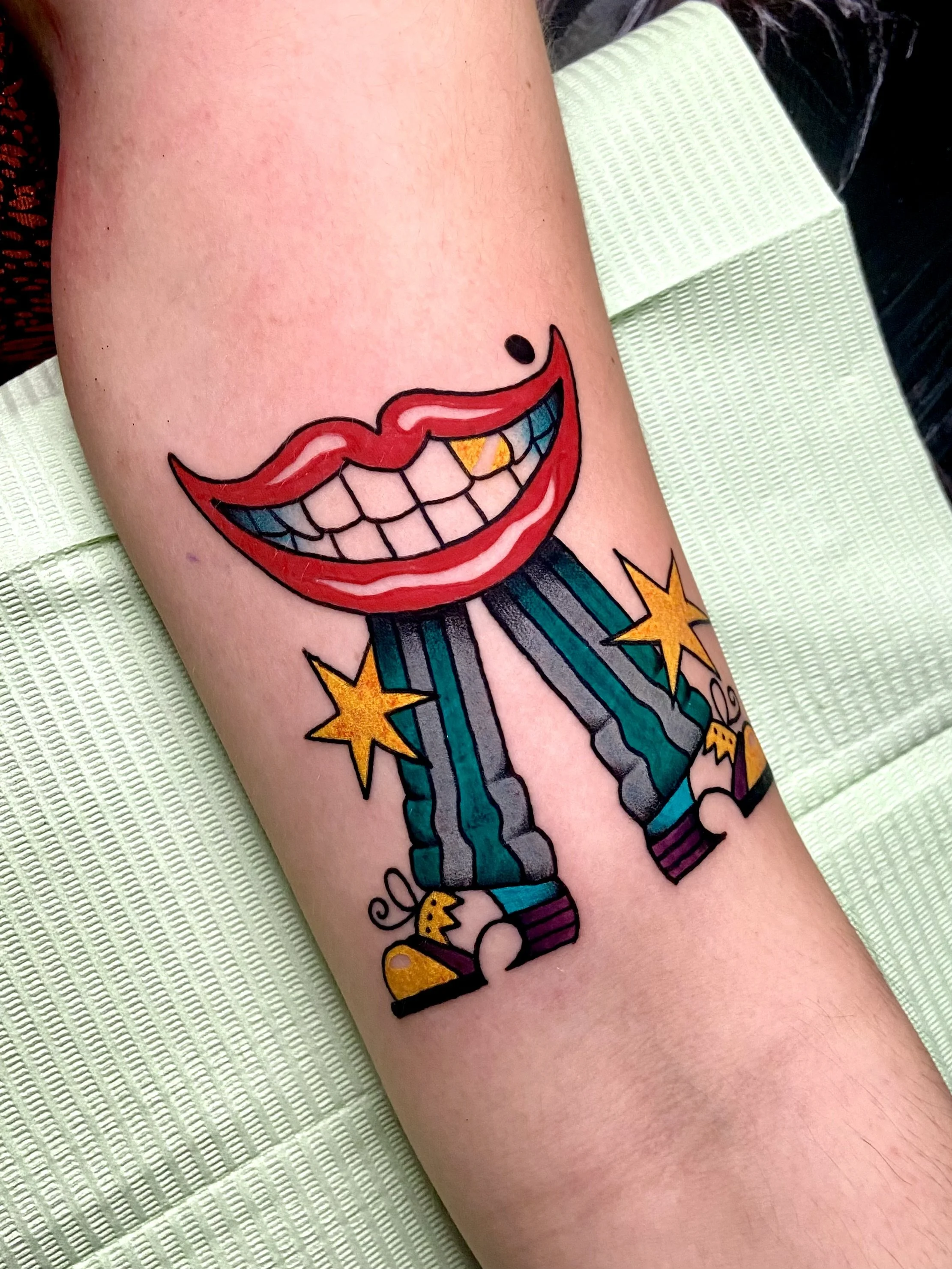 Tattoo of a cartoon red mouth wearing green and black striped pants, yellow stars, and small shoes.