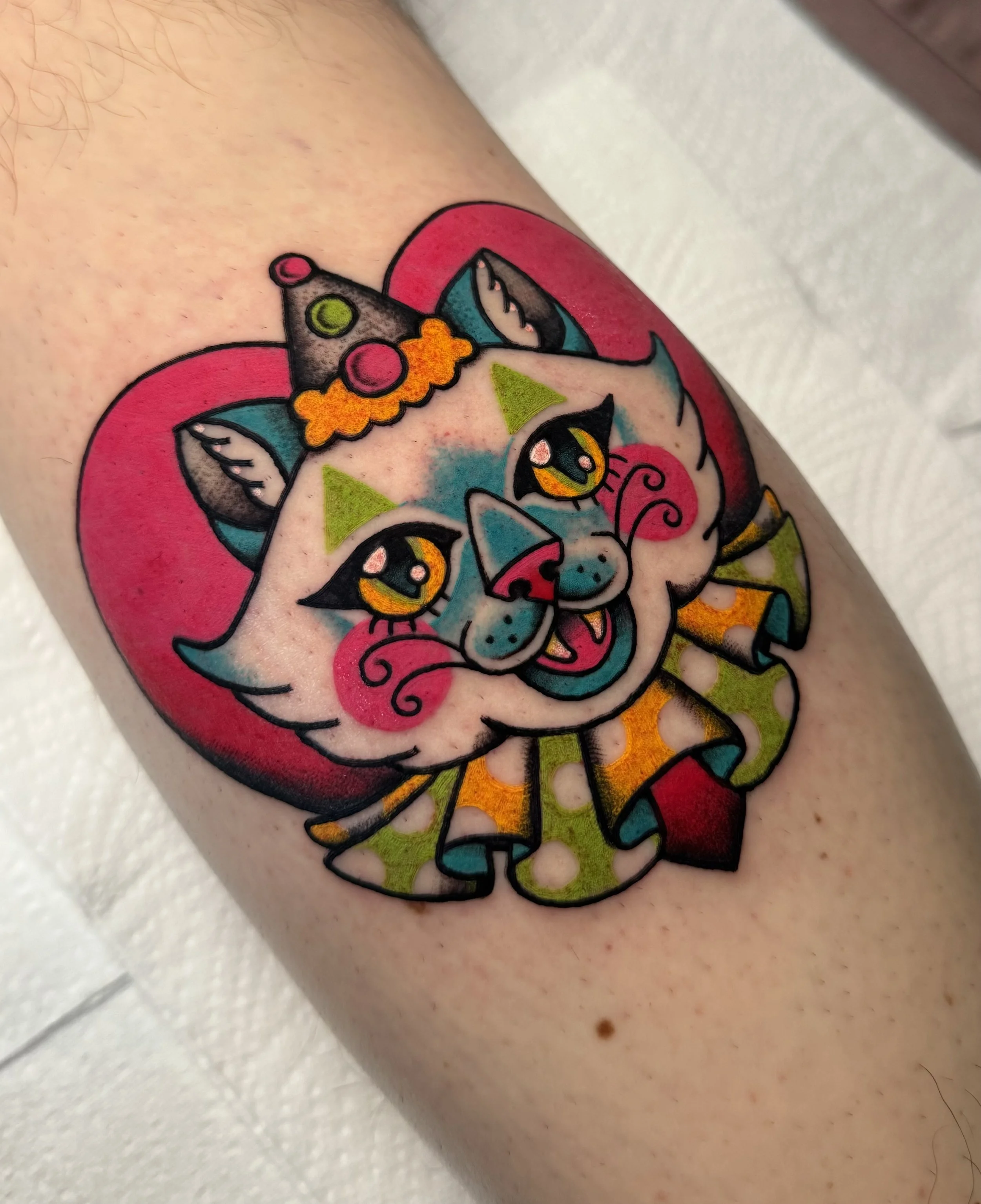 Colorful cartoon-style tattoo of a cute pink and white cat with large yellow eyes, wearing a clown hat, with a pink background.