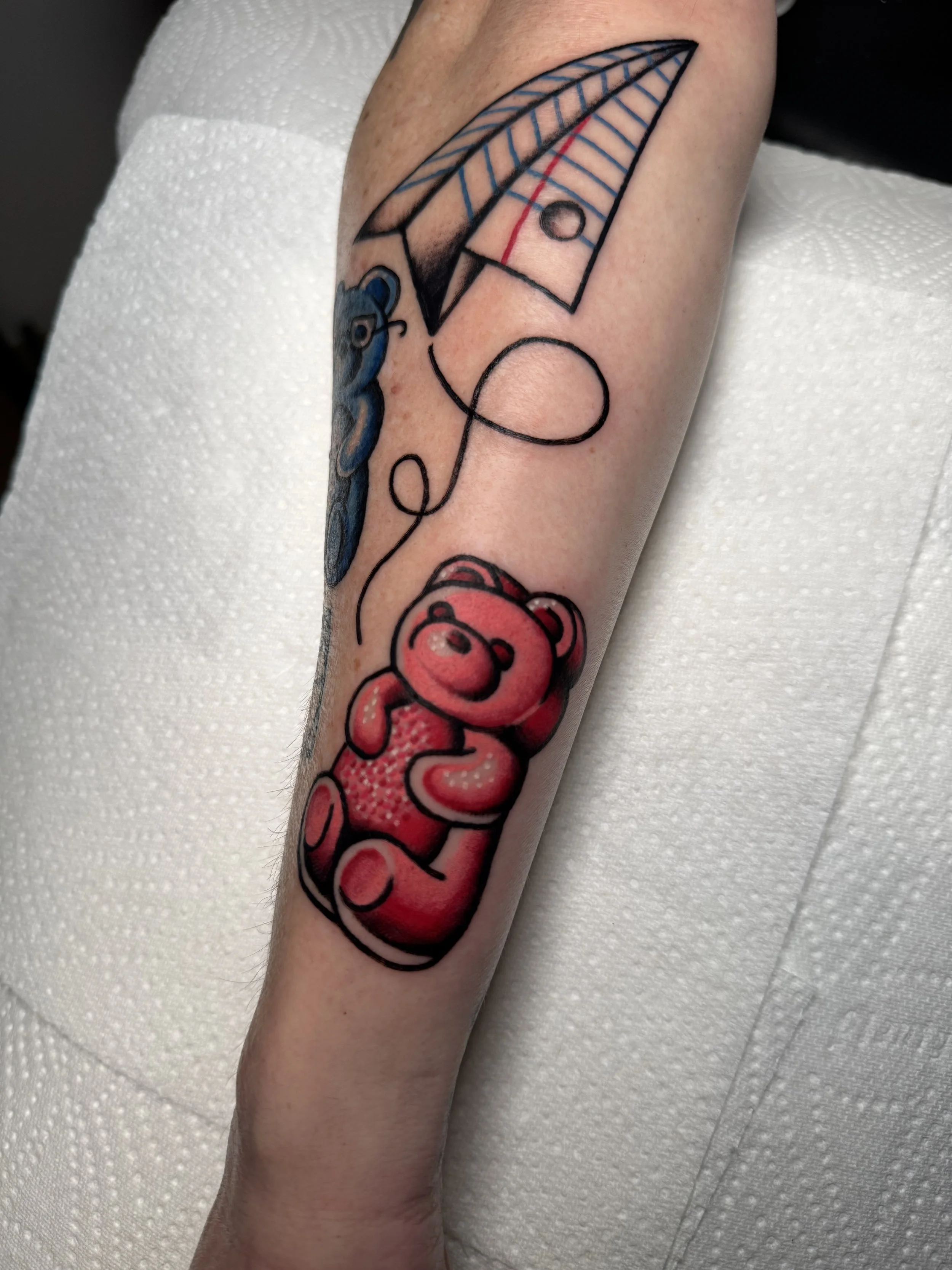 Tattoo of a red teddy bear, a blue bird, and a paper airplane on a person's arm.