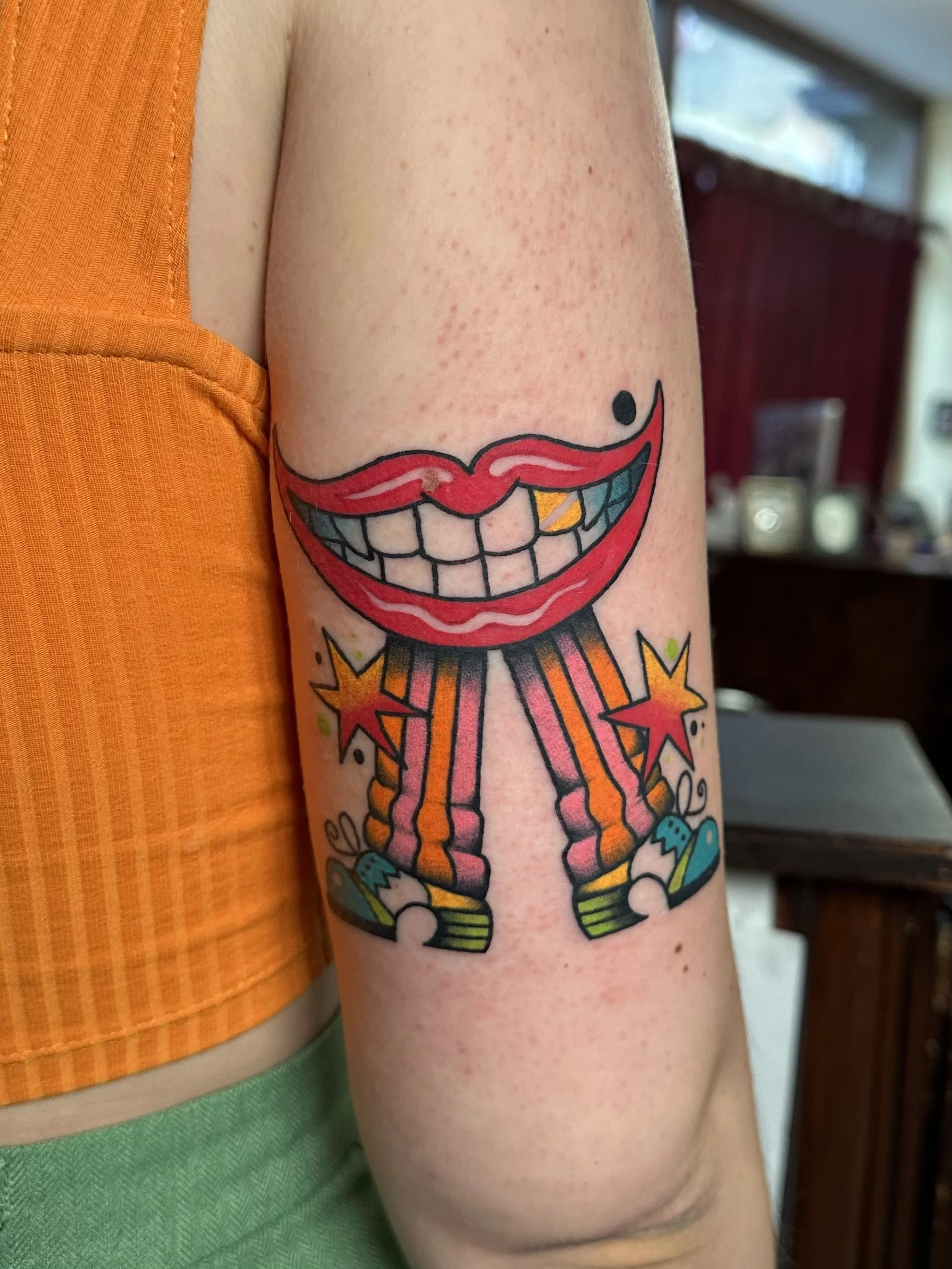 Tattoo of a grinning cartoonish mouth with teeth, a dartboard, and sneakers on a person's upper arm.