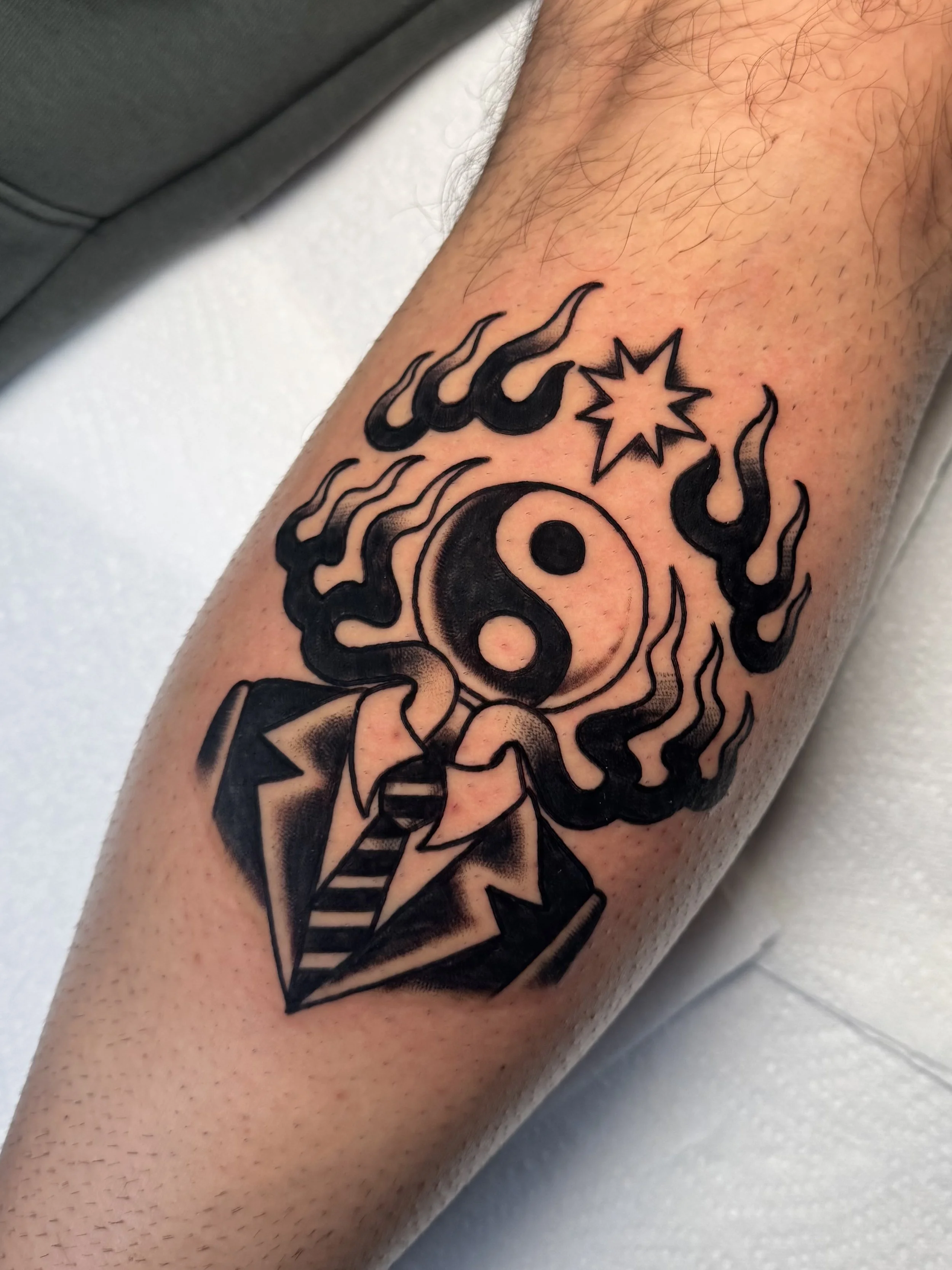 Tattoo of a stylized yin-yang symbol surrounded by flames, a star, and a geometric pattern on a person's forearm.