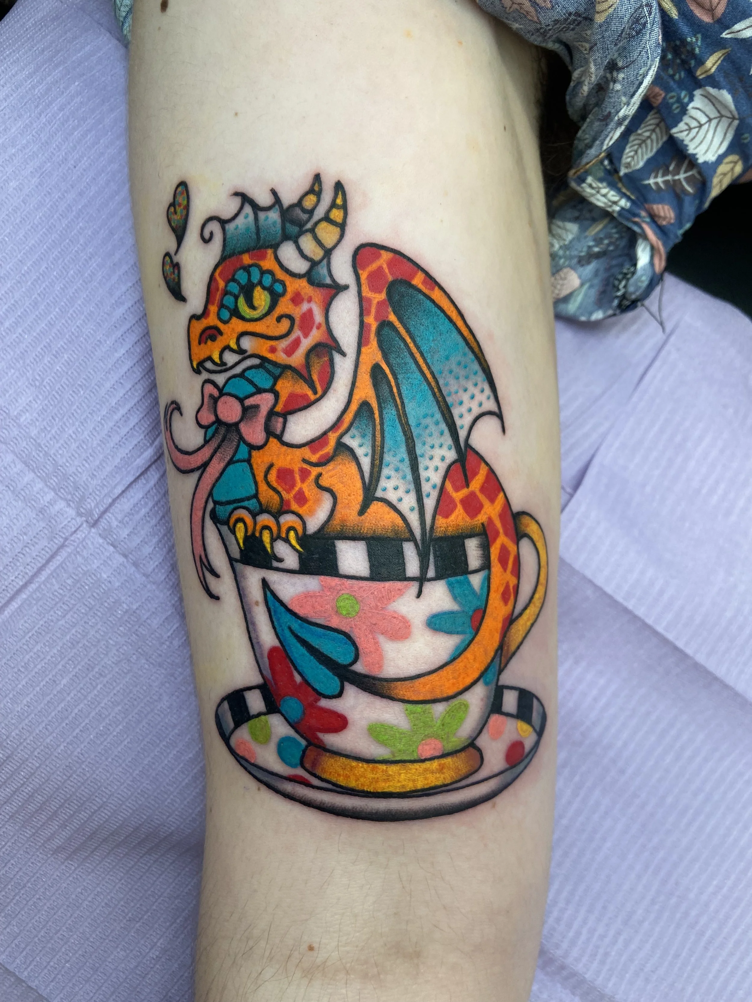 Colorful tattoo of a cartoon-style dragon sitting inside a decorative teacup.