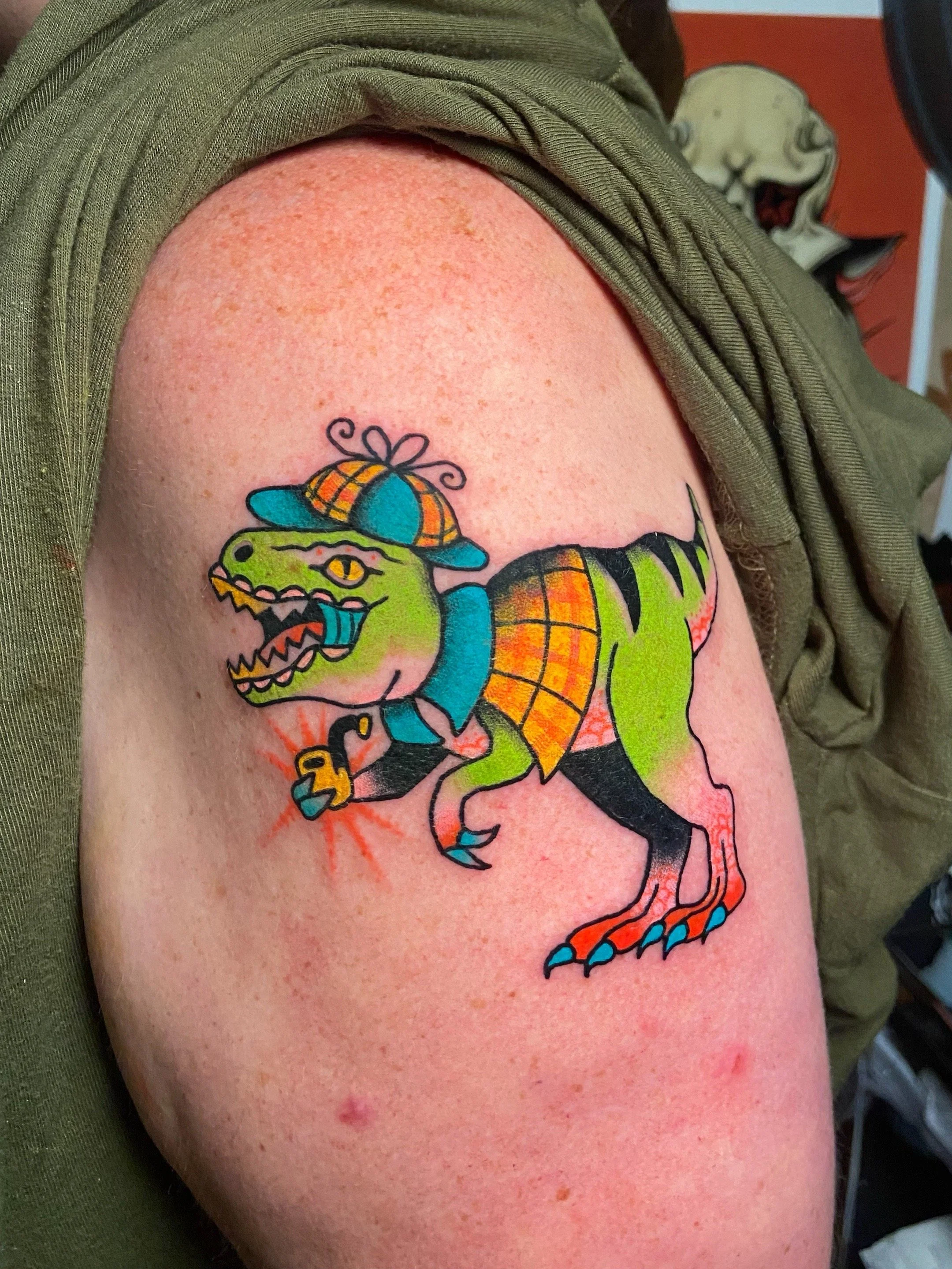 Colorful tattoo of a dinosaur wearing a plaid hat, a scarf, and holding a small yellow smoking pipe. The dinosaur is green with black stripes and orange claws, depicted in a cartoon style. It is a T-rex made to look like Sherlock Homes.