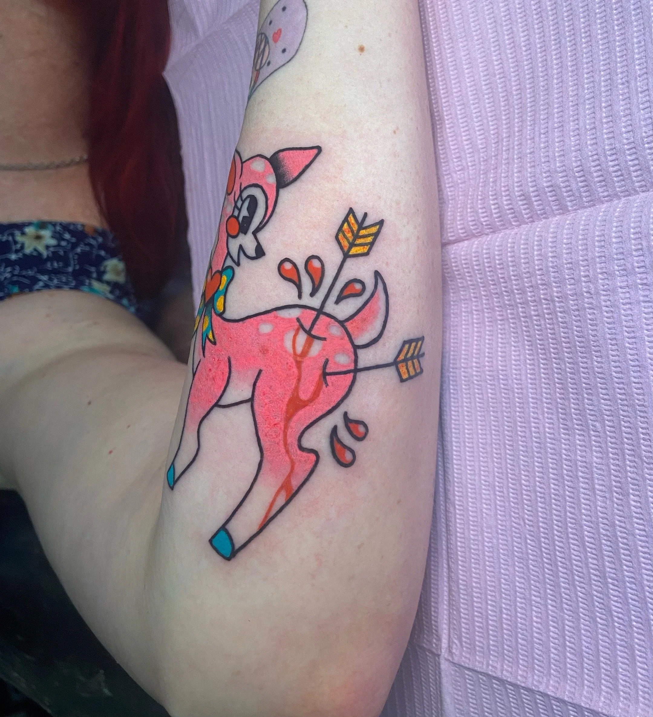 Colorful tattoo of a pink cartoon deer with multiple arrows and some red droplets on the person's upper arm.
