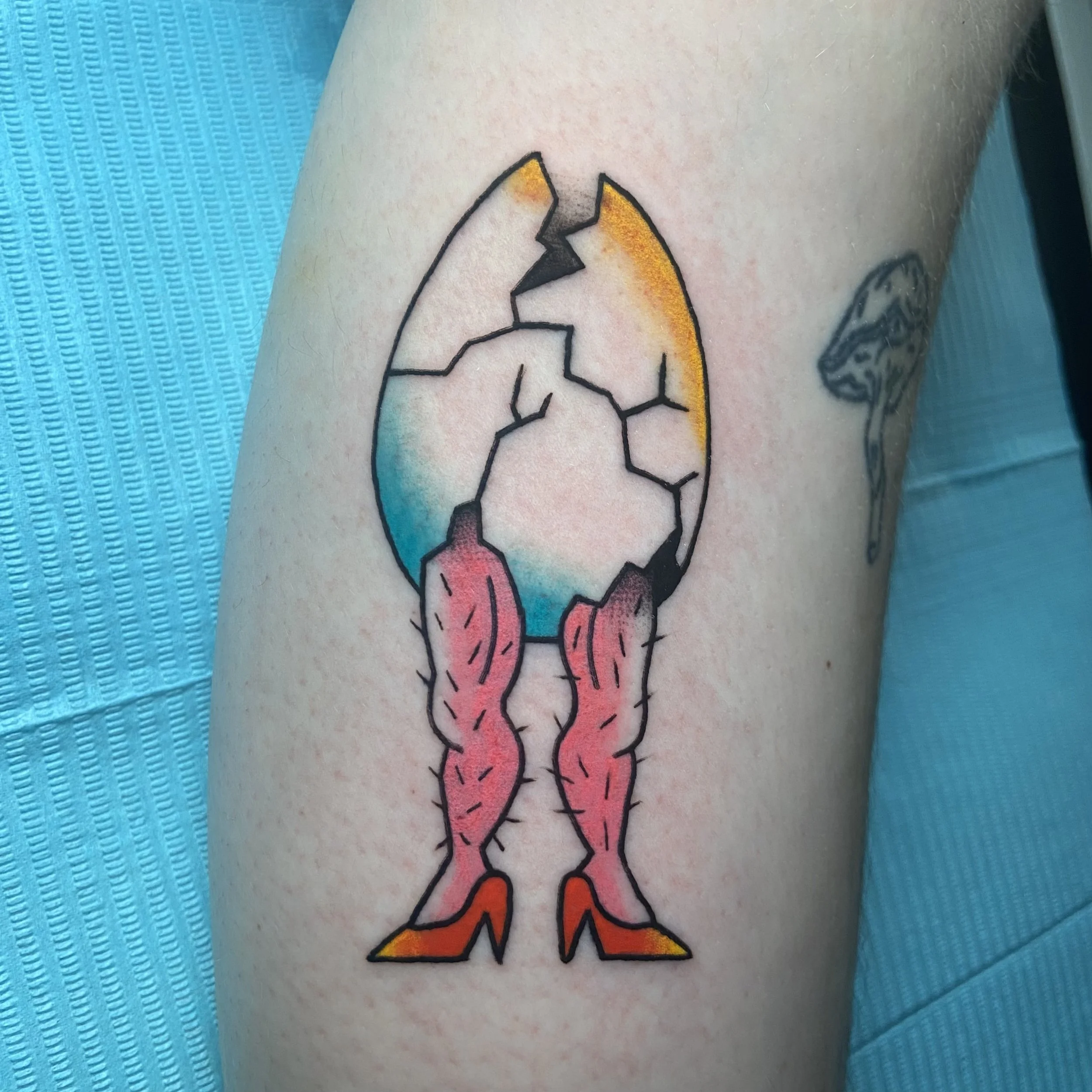 A colorful tattoo of a cracked eggshell with pink hairy legs and red high heels.