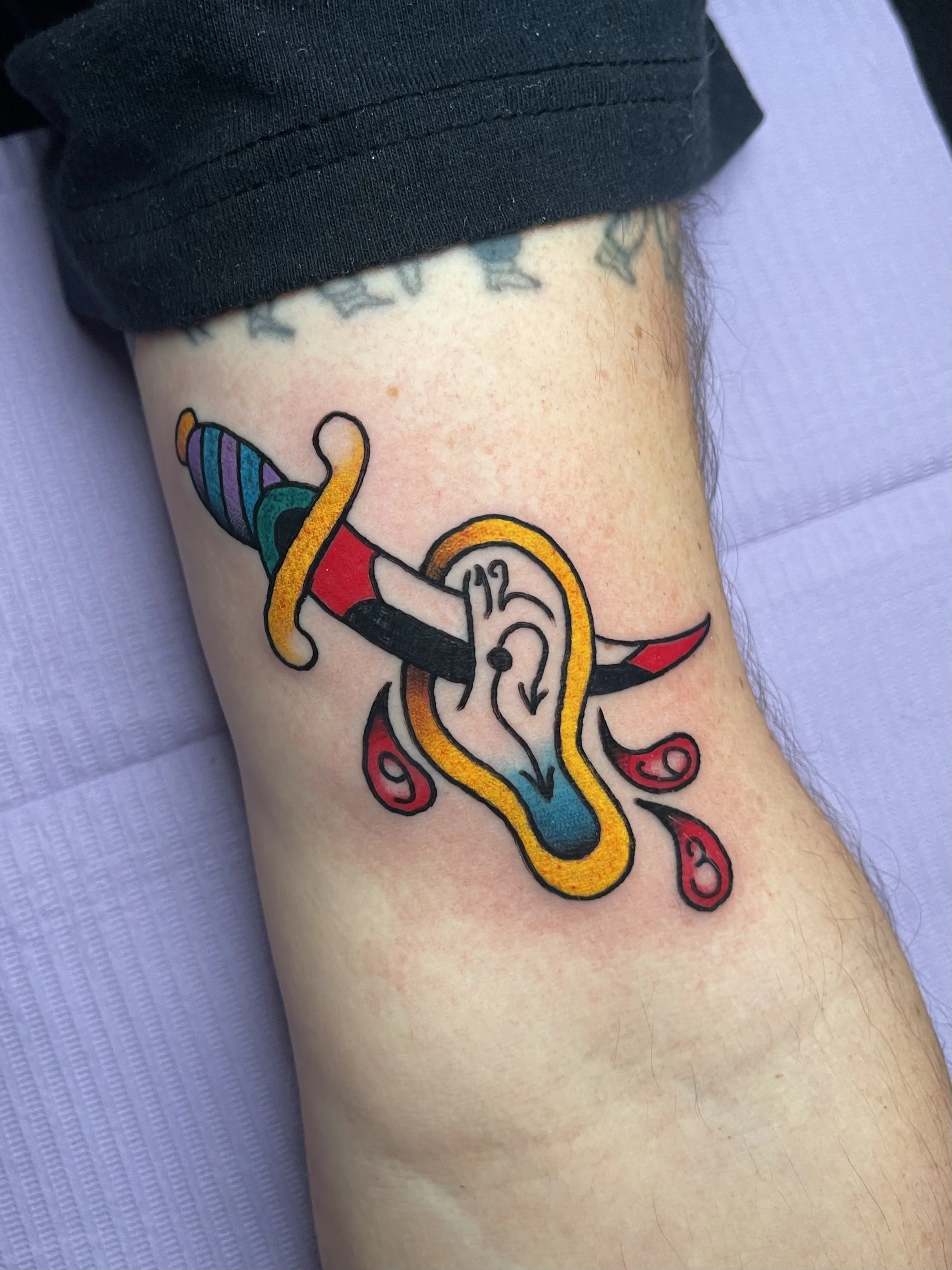 Colorful tattoo of a sword and a Dali inspired clock on a person's arm, with red decorative elements and a dagger going through it.