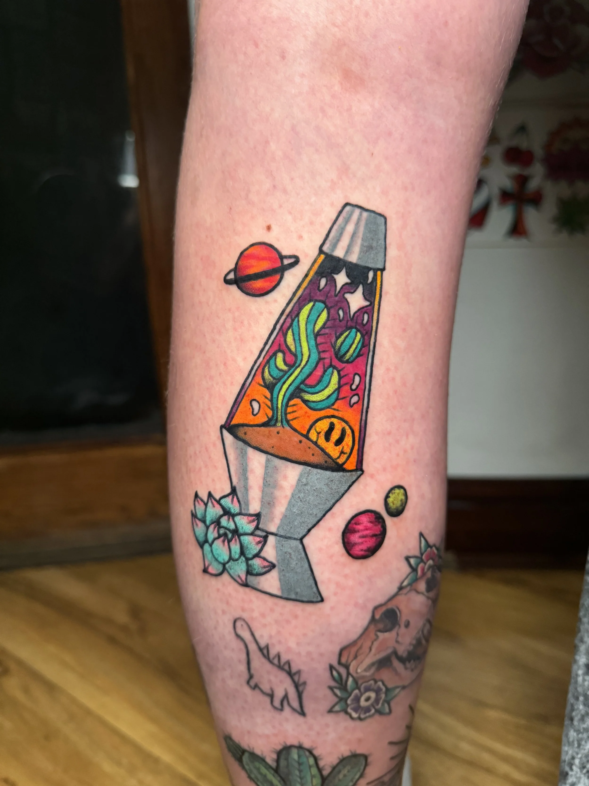 Colorful tattoo of a lava lamp with liquid and abstract shapes inside, surrounded by planets, a cactus, a skull, and other decorative elements.