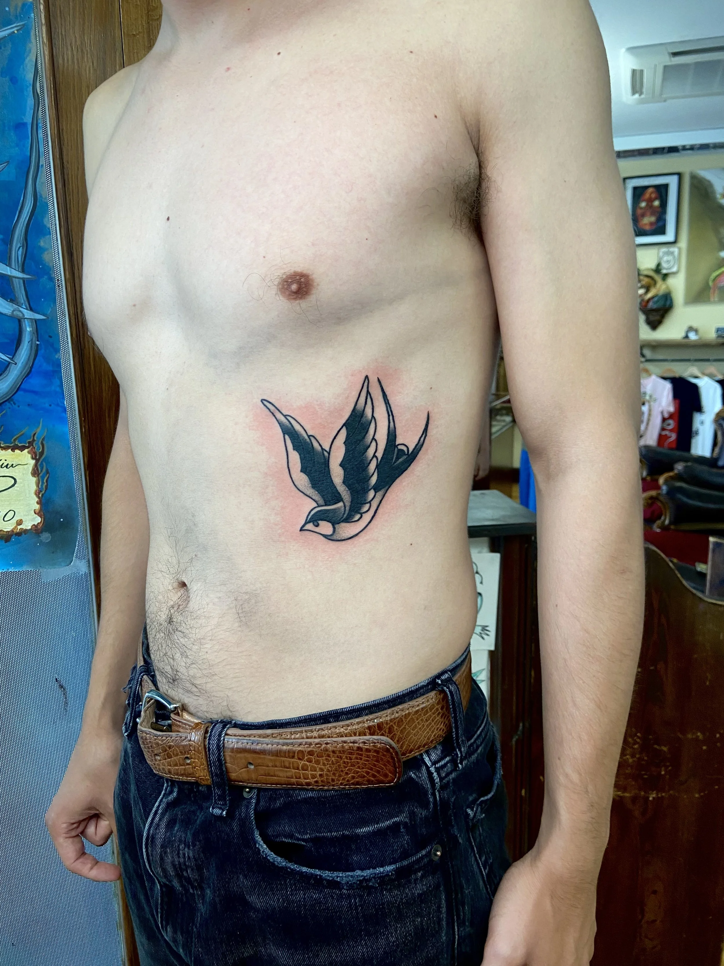 A person with a swallow tattoo on their ribs light skin, and wearing dark jeans with a brown belt, standing in what appears to be a shop or tattoo parlour.
