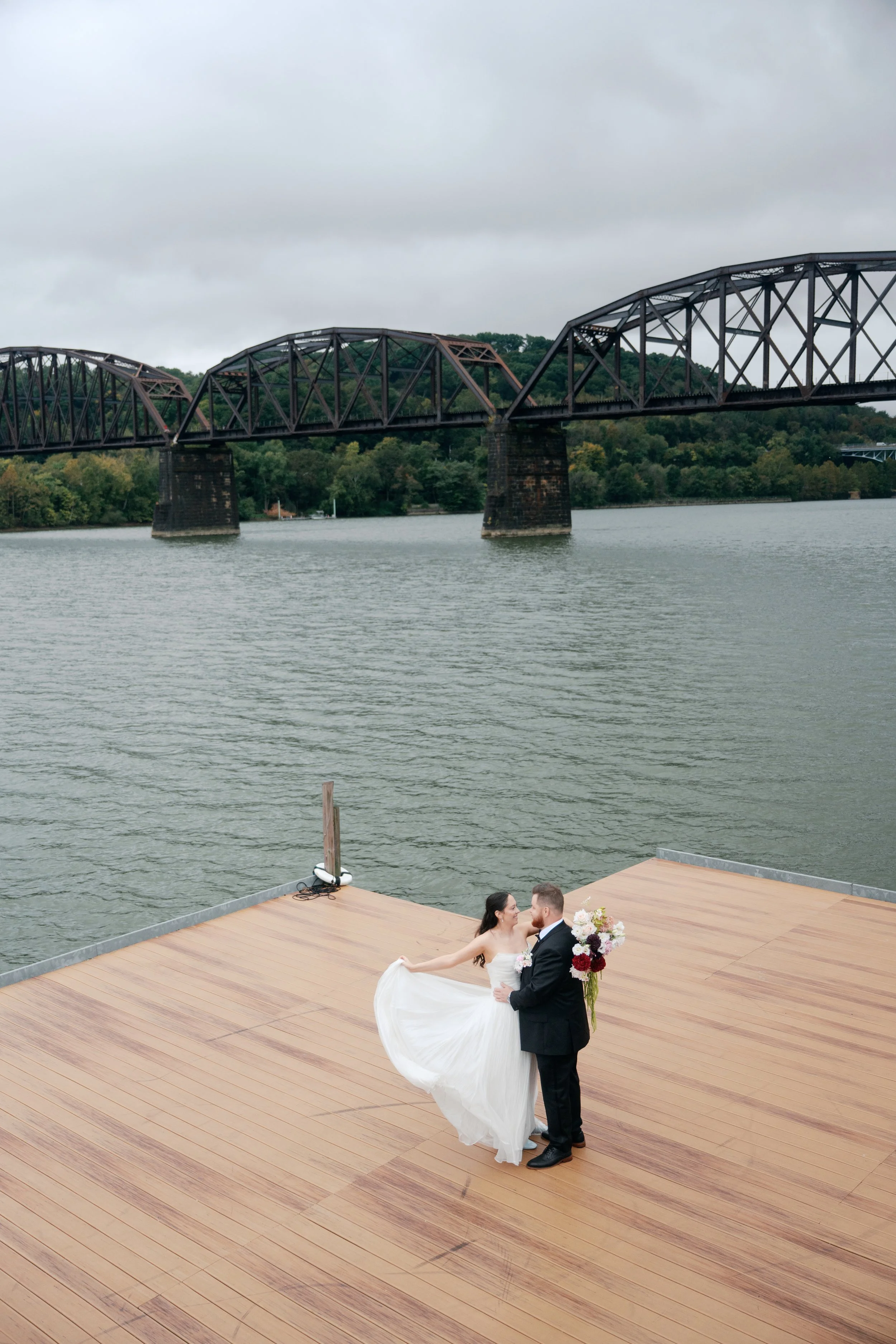 A romantic waterfront wedding portrait featuring a cascading burgundy and blush bridal bouquet, a coordinating groom’s floral pocket arrangement, and a flowing white bridal gown. Soft pink and ivory flowers contrast with rich wine-colored blooms and 