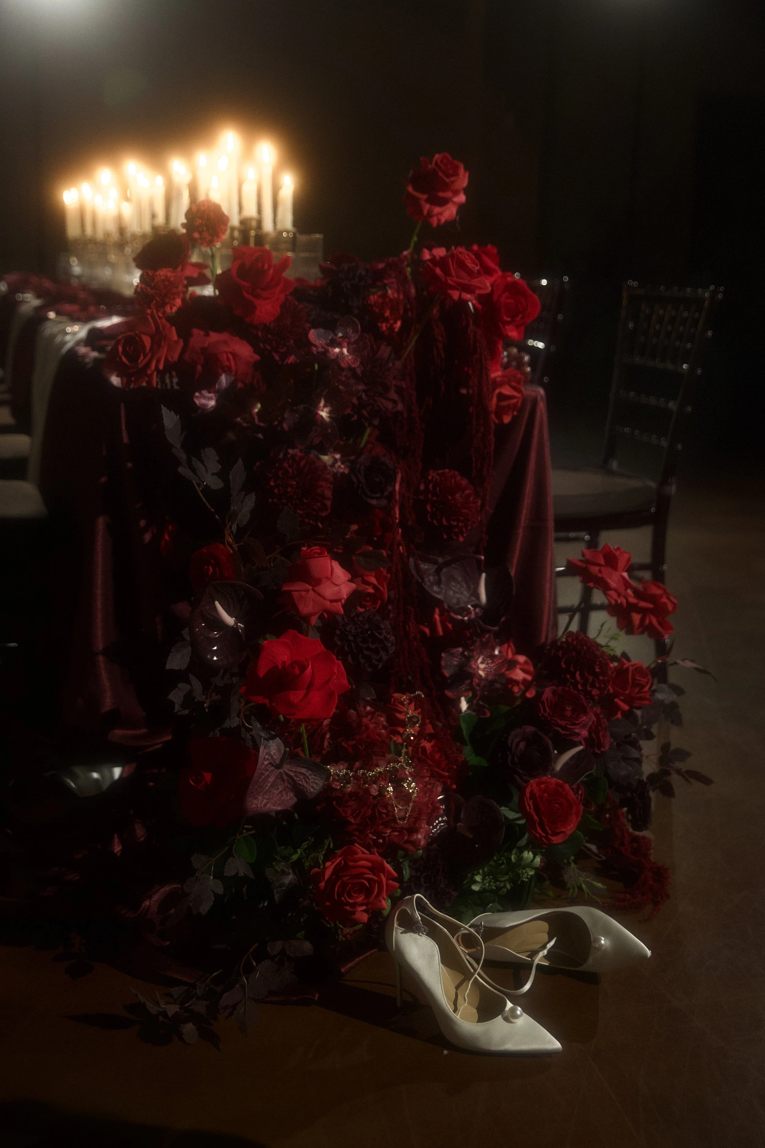 A table decorated with red flowers and candles, with a pair of white high-heeled shoes placed underneath.