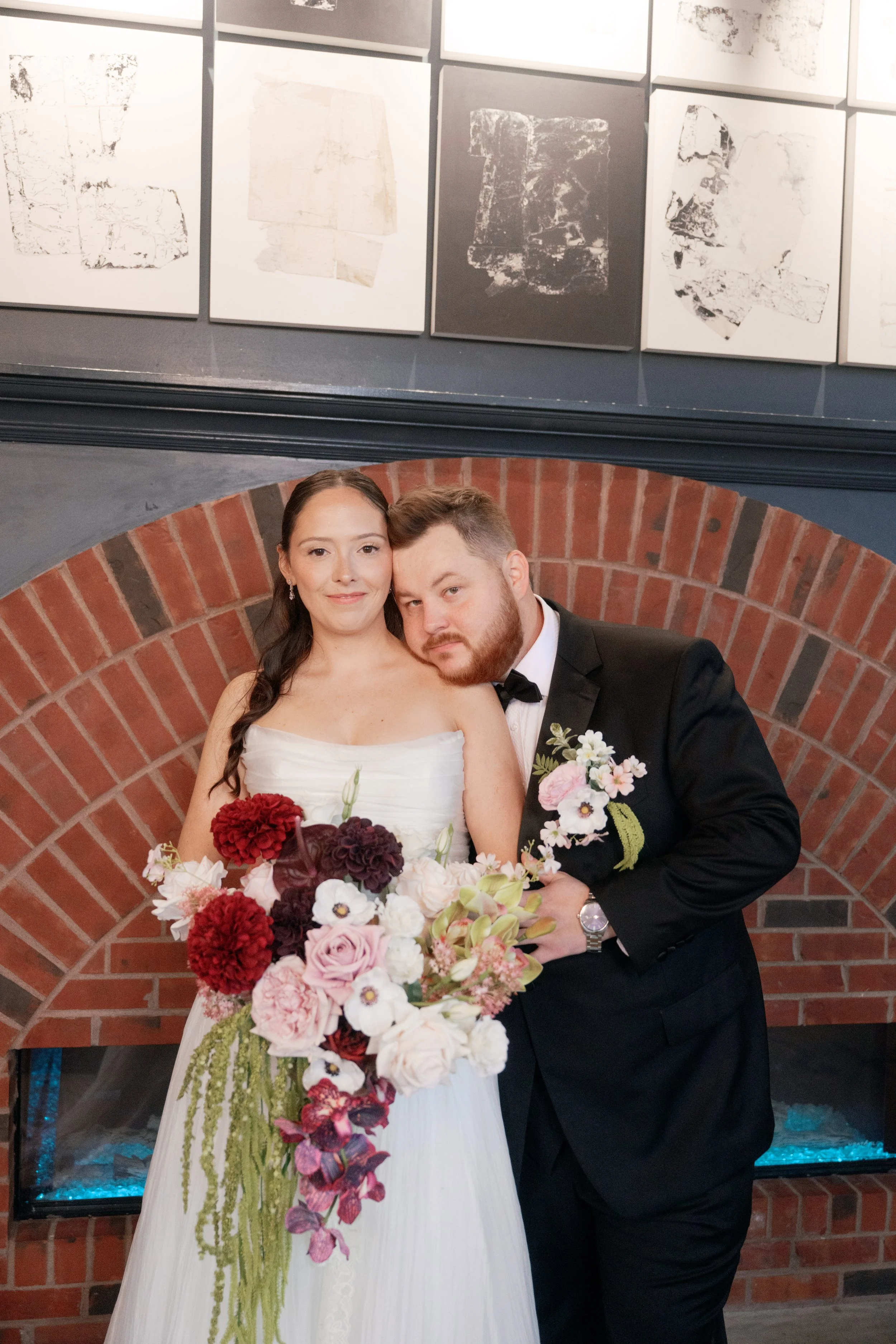 A cascading bridal bouquet in blush pink, ivory, burgundy, and soft green brings romantic texture to this wedding portrait. Sculptural blooms, trailing green amaranthus, and cascading orchids complement the groom’s coordinating floral pocket arrangem