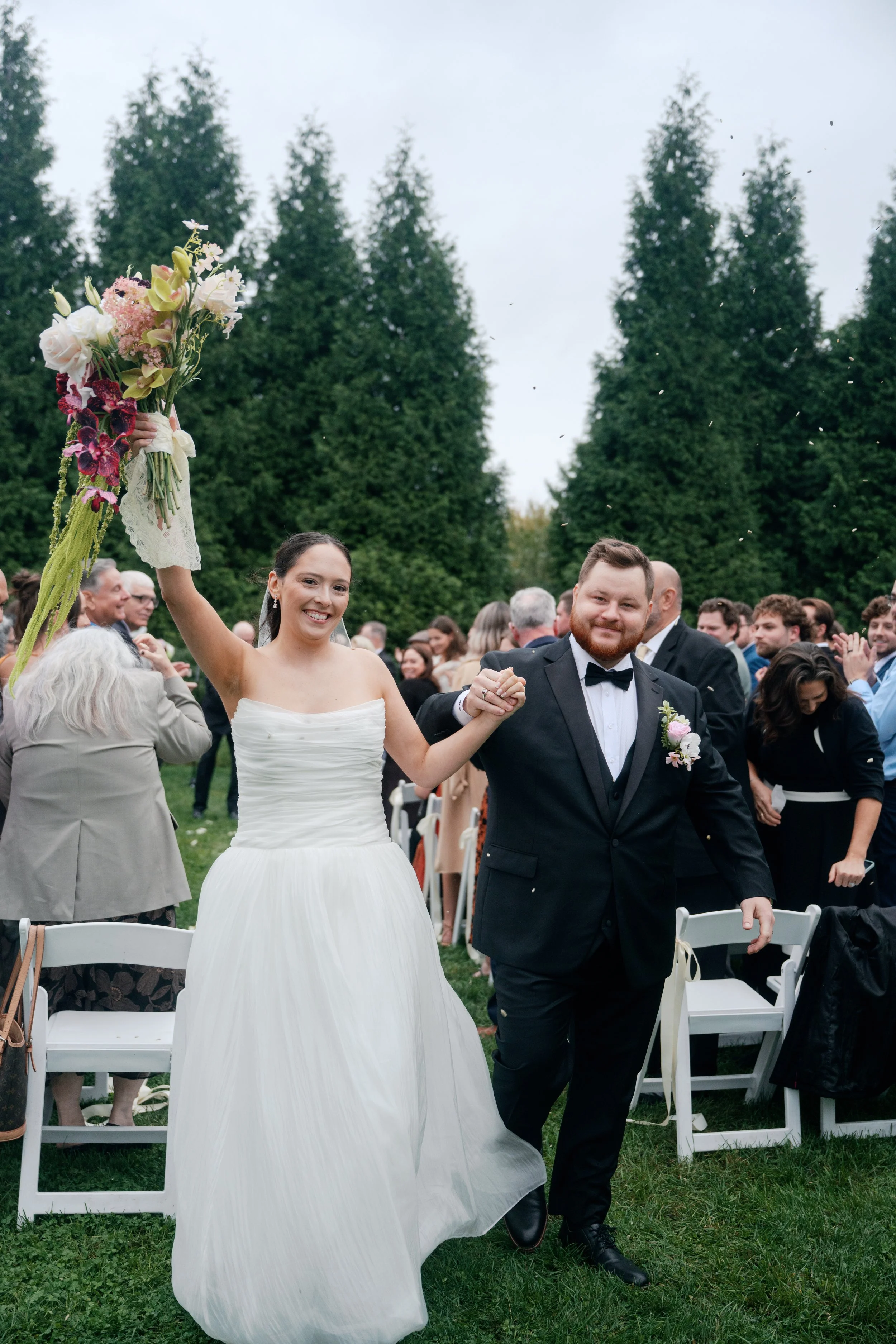 A newlywed couple holding hands and smiling, walking outdoors at a wedding reception. The bride is holding a bouquet of flowers, and the groom is dressed in a black tuxedo with a bow tie. There are guests in the background, seated and standing, with 