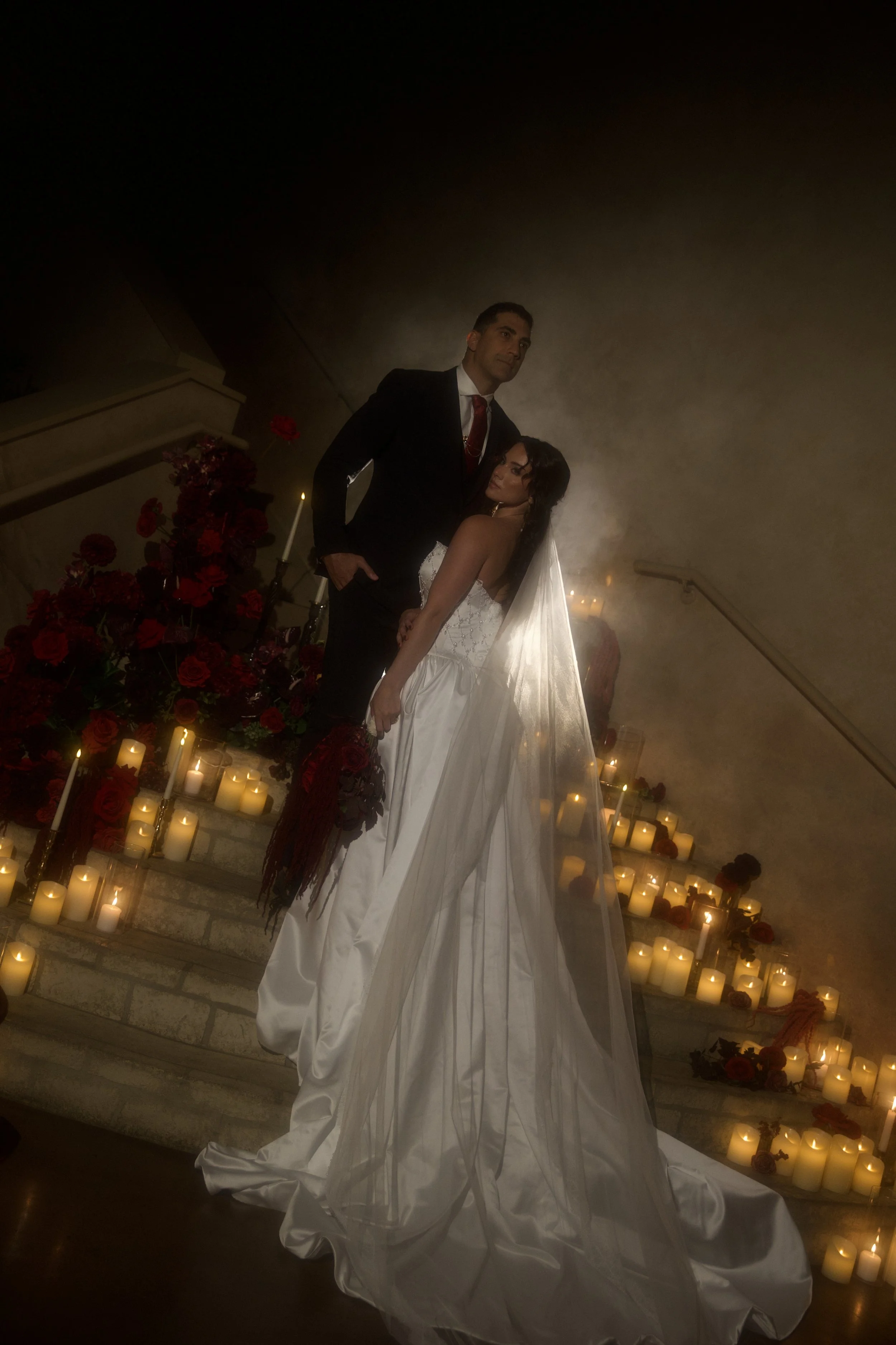 Dark romance wedding inspiration at Bella Sera Event Villa near Pittsburgh, featuring a cascading red and burgundy bridal bouquet and a candlelit staircase filled with dramatic floral arrangements. Floral design by S.R. Event Design & Decor pairs ric