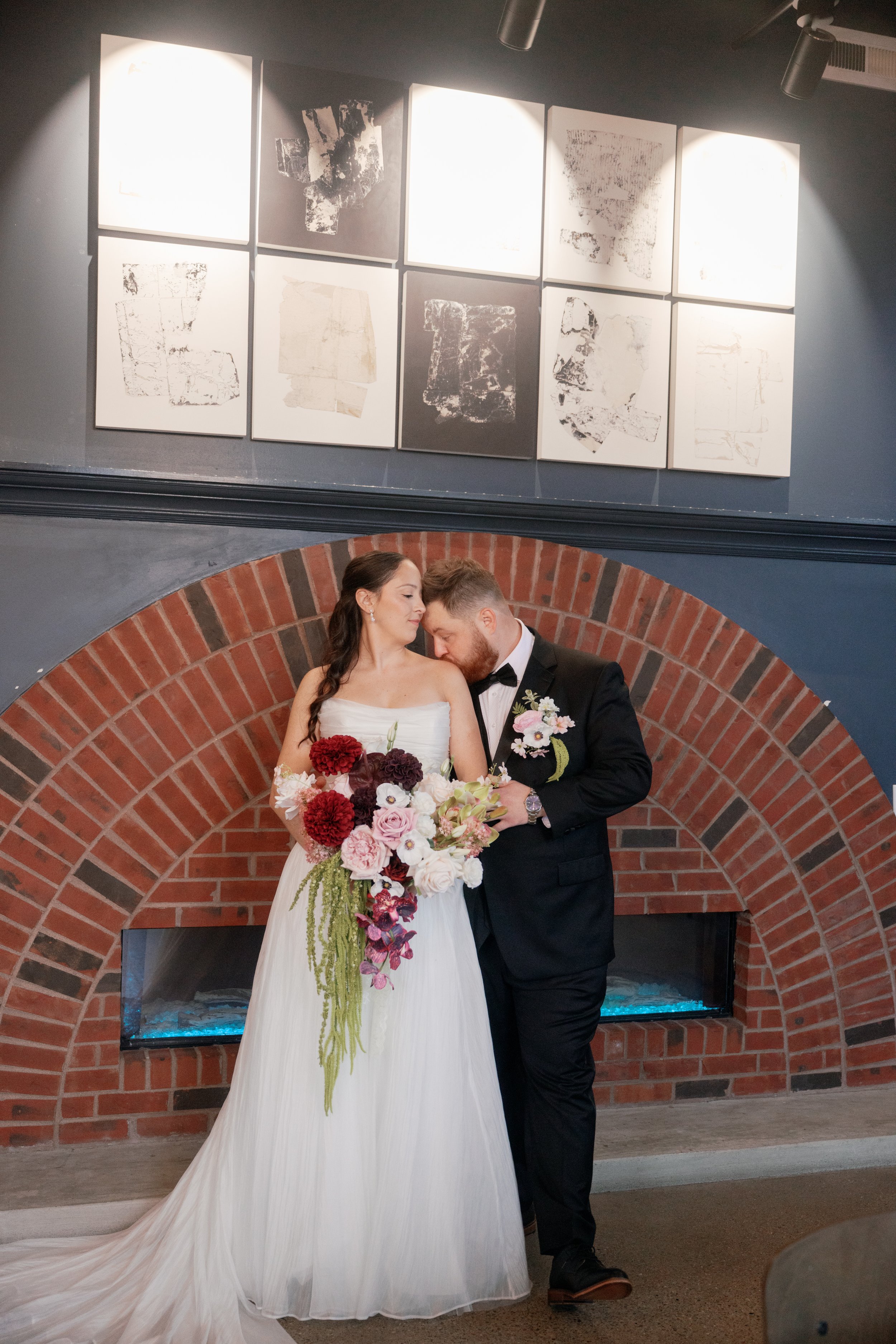 Bride and groom leaning together beside a brick fireplace, with a large blush and burgundy bouquet cascading down the bride’s white gown. @TKPhotoand Film 
Planner : @MadilineKellyevents 
Florist @SREventDesign & Decor 