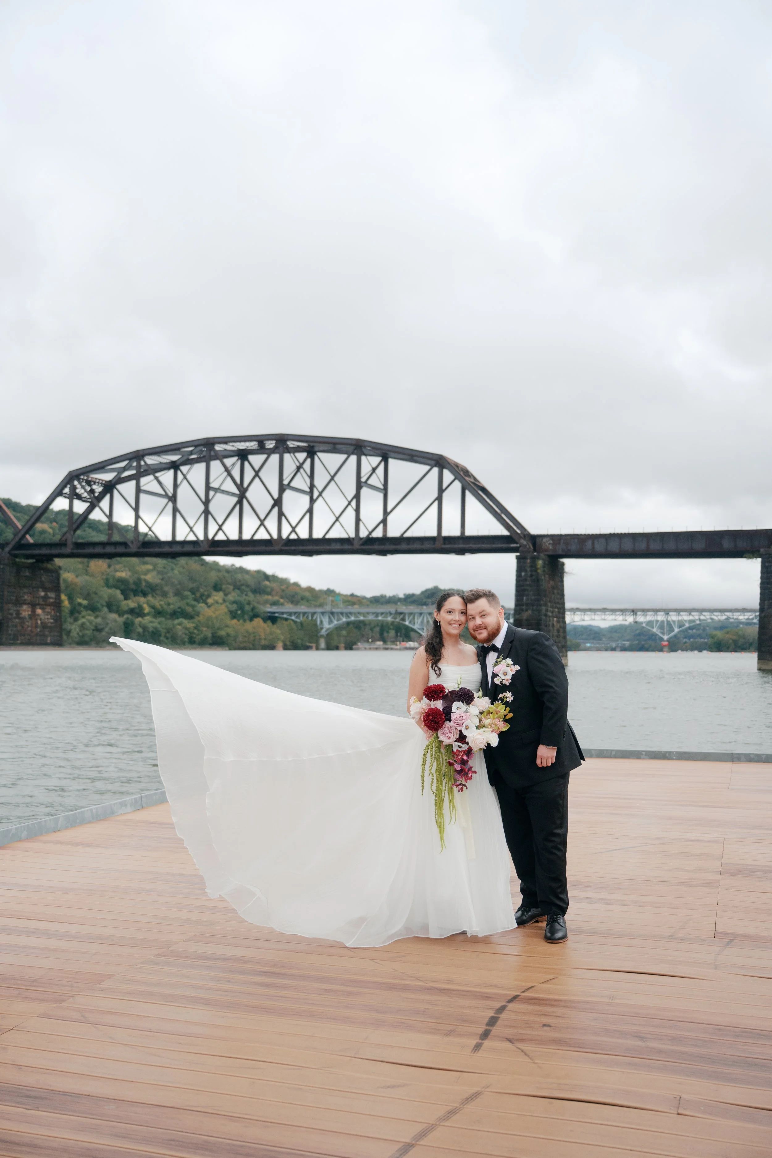 A romantic waterfront wedding portrait featuring a cascading burgundy and blush bridal bouquet, a coordinating groom’s floral pocket arrangement, and a flowing white bridal gown. Soft pink and ivory flowers contrast with rich wine-colored blooms and 