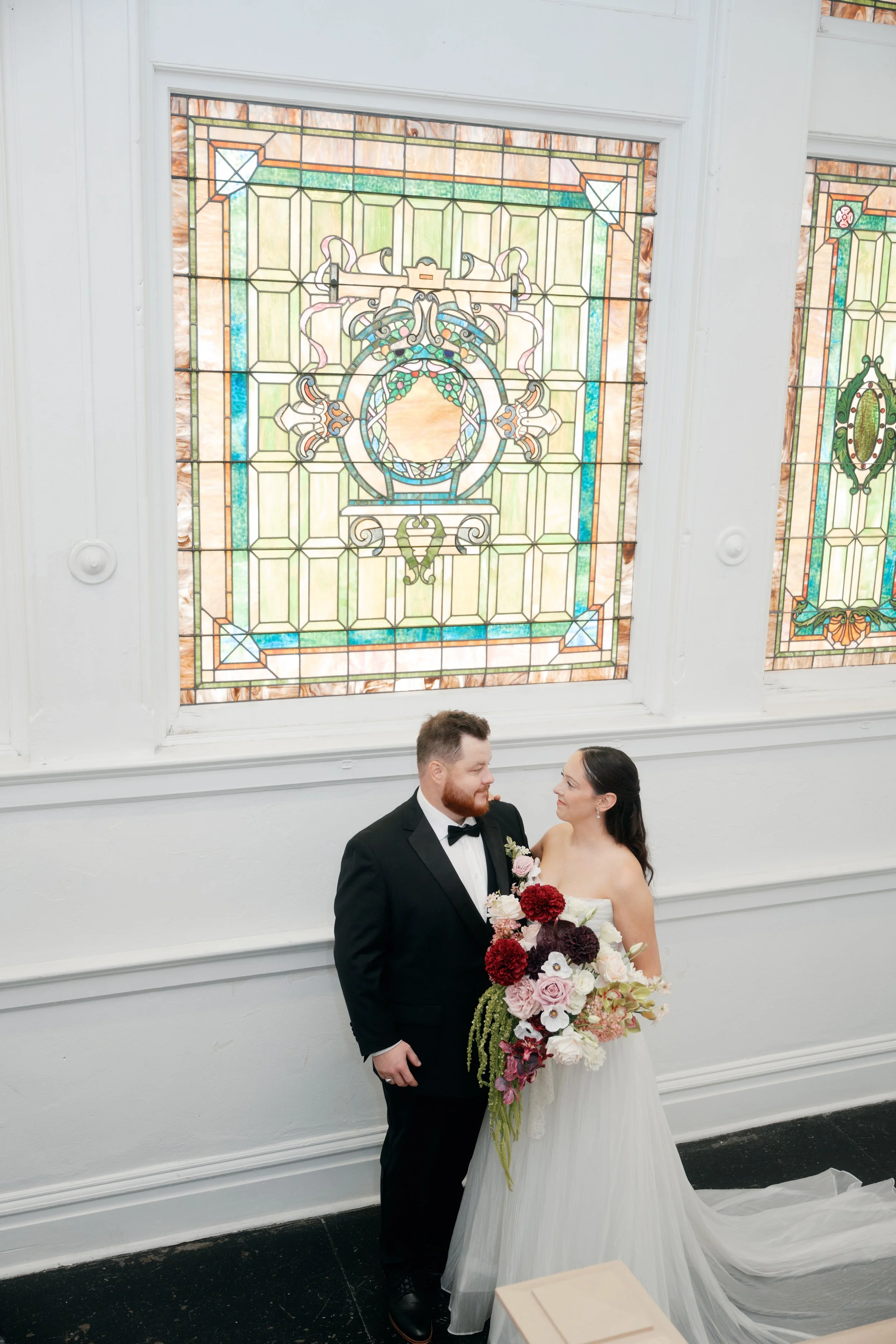 A romantic portrait featuring a cascading burgundy and blush bridal bouquet, a coordinating groom’s floral pocket arrangement, and a flowing white bridal gown. Soft pink and ivory flowers contrast with rich wine-colored blooms and trailing greenery. 