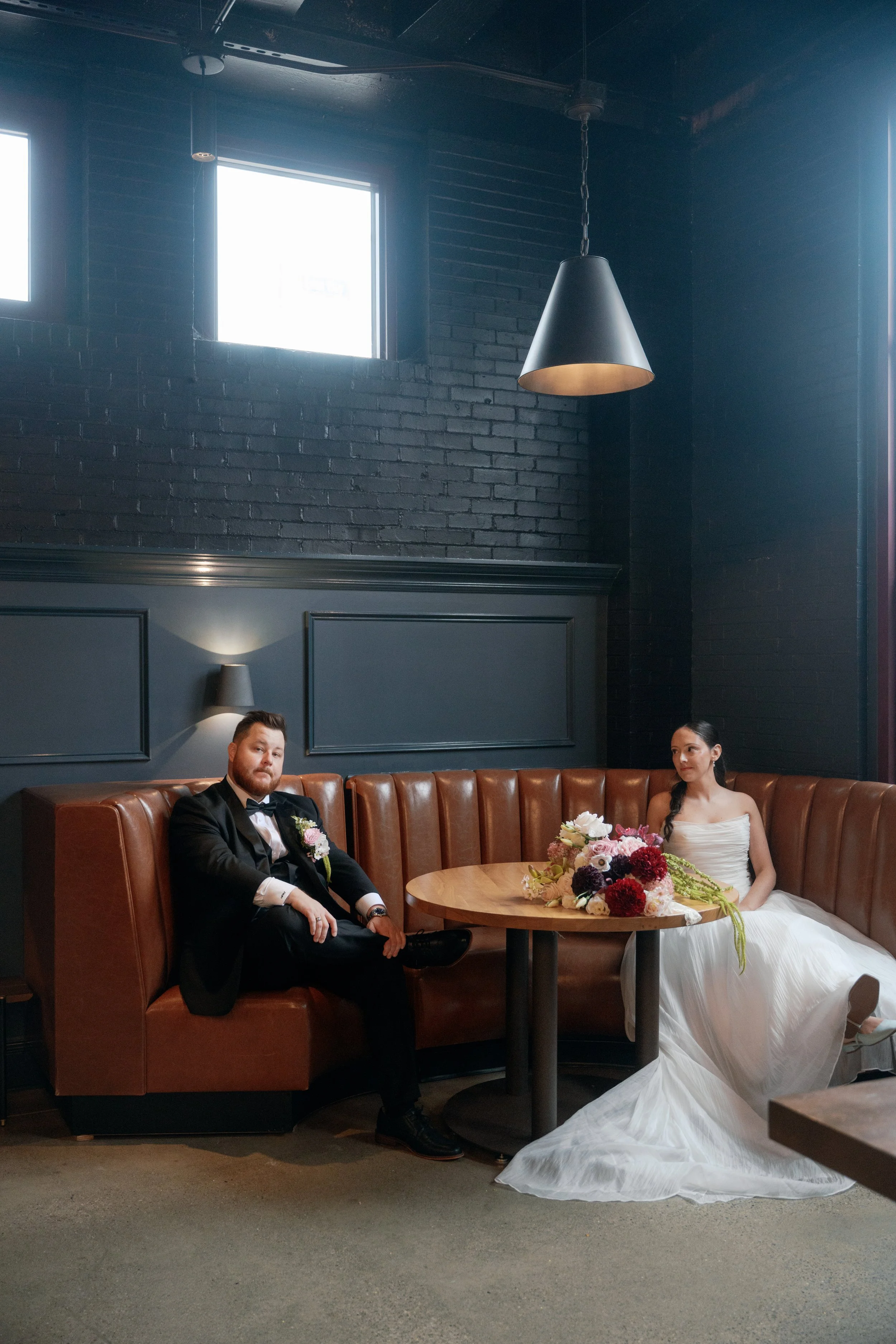 Modern wedding style meets romantic floral design in this relaxed couple’s portrait, featuring a cascading blush and burgundy bridal bouquet resting on a café table. Ivory blooms and trailing greenery soften the dark walls and caramel leather seating