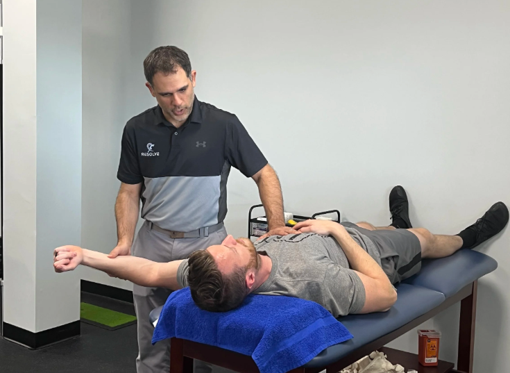 A man lies on a treatment table, stretching his arm, while a healthcare professional checks his shoulder in a clinical setting.