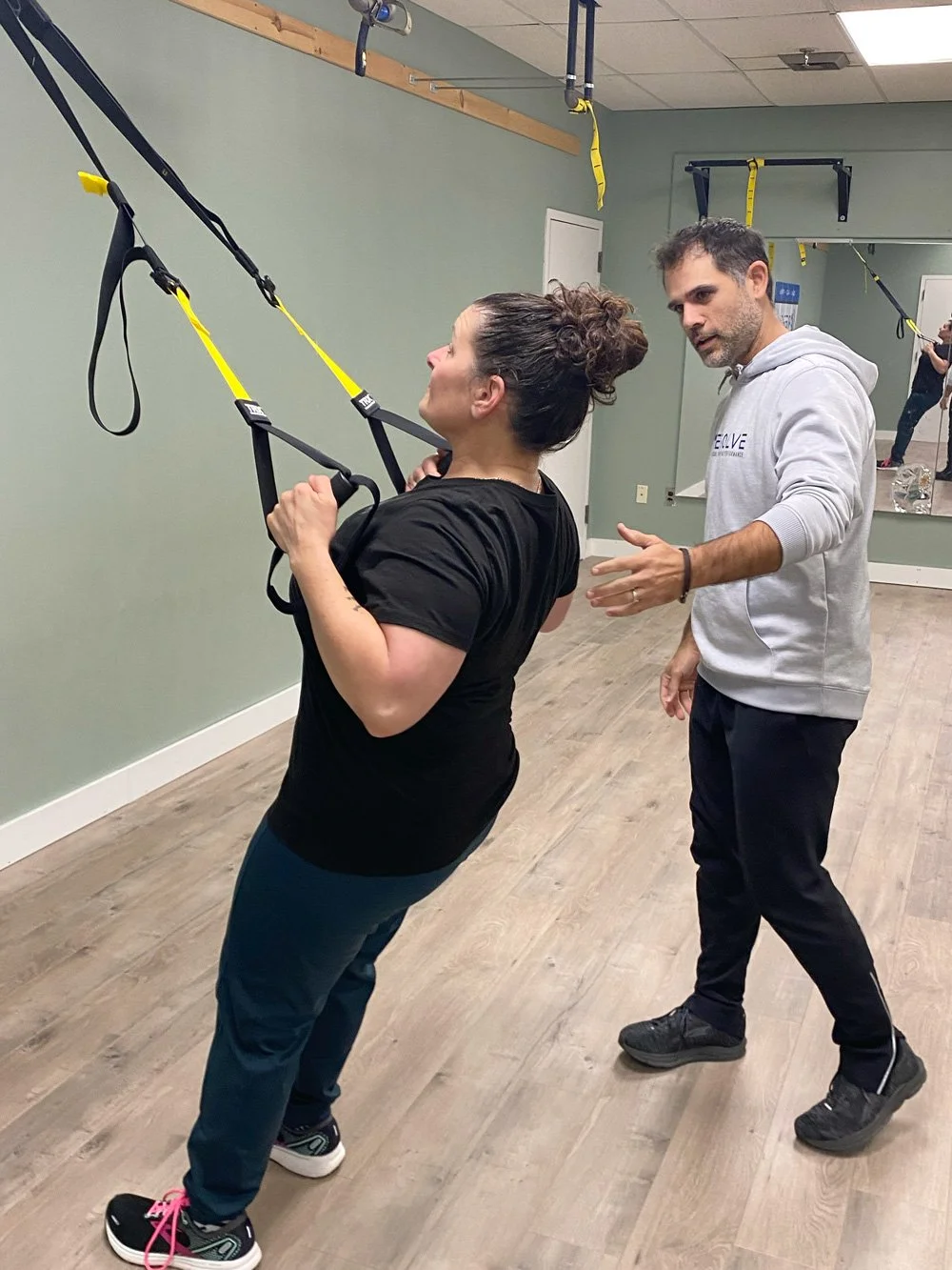 A woman doing suspension training exercises with a trainer guiding her in a gym or fitness studio.