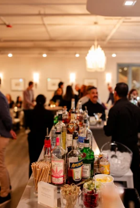 A bar setup at a social event with various bottles of alcohol, drinks, snacks, and people in the background in a well-lit room.