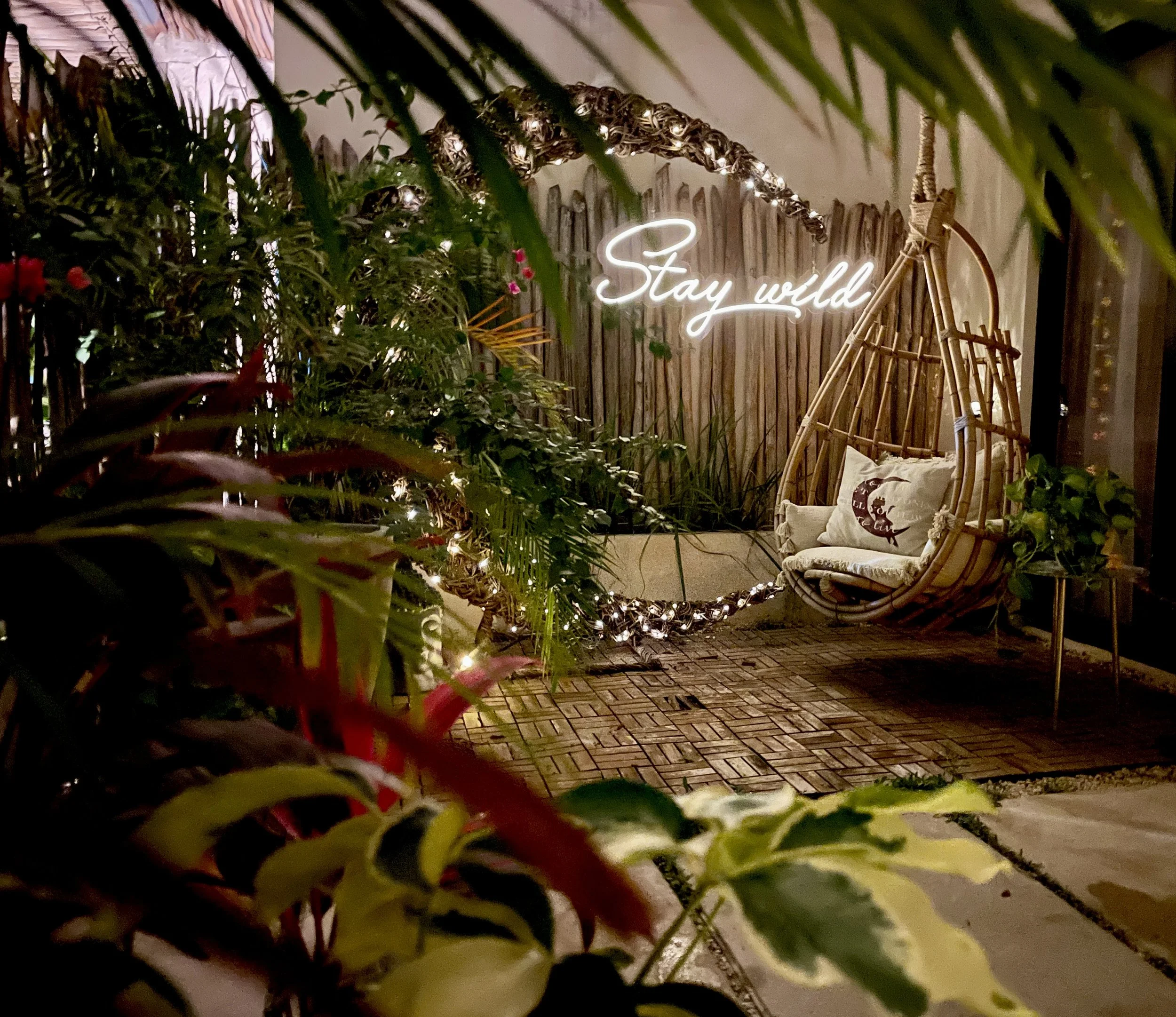 A cozy outdoor seating area with a rattan hanging swing chair containing green cushions and a pillow, a small bench with plants, a wooden fence backdrop, and a neon sign that reads 'Stay wild' illuminated in white.
