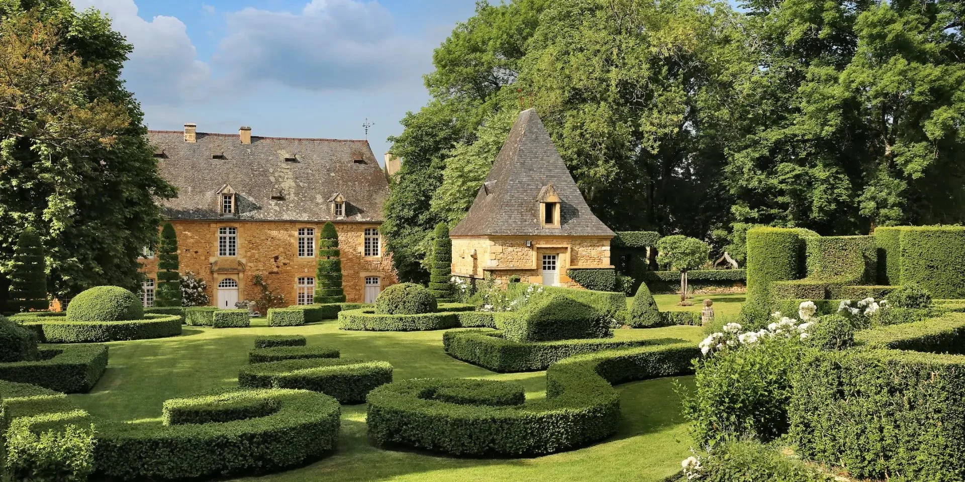 A stone manor house with steep rooflines surrounded by a well-manicured formal garden with trimmed hedges, topiary shapes, and flowering plants