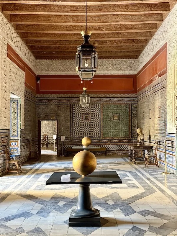 Interior of a museum or historic house with decorative patterned walls, wooden ceiling, hanging lanterns, and display of sculptures and furniture, including a bust and a large round object on a pedestal.