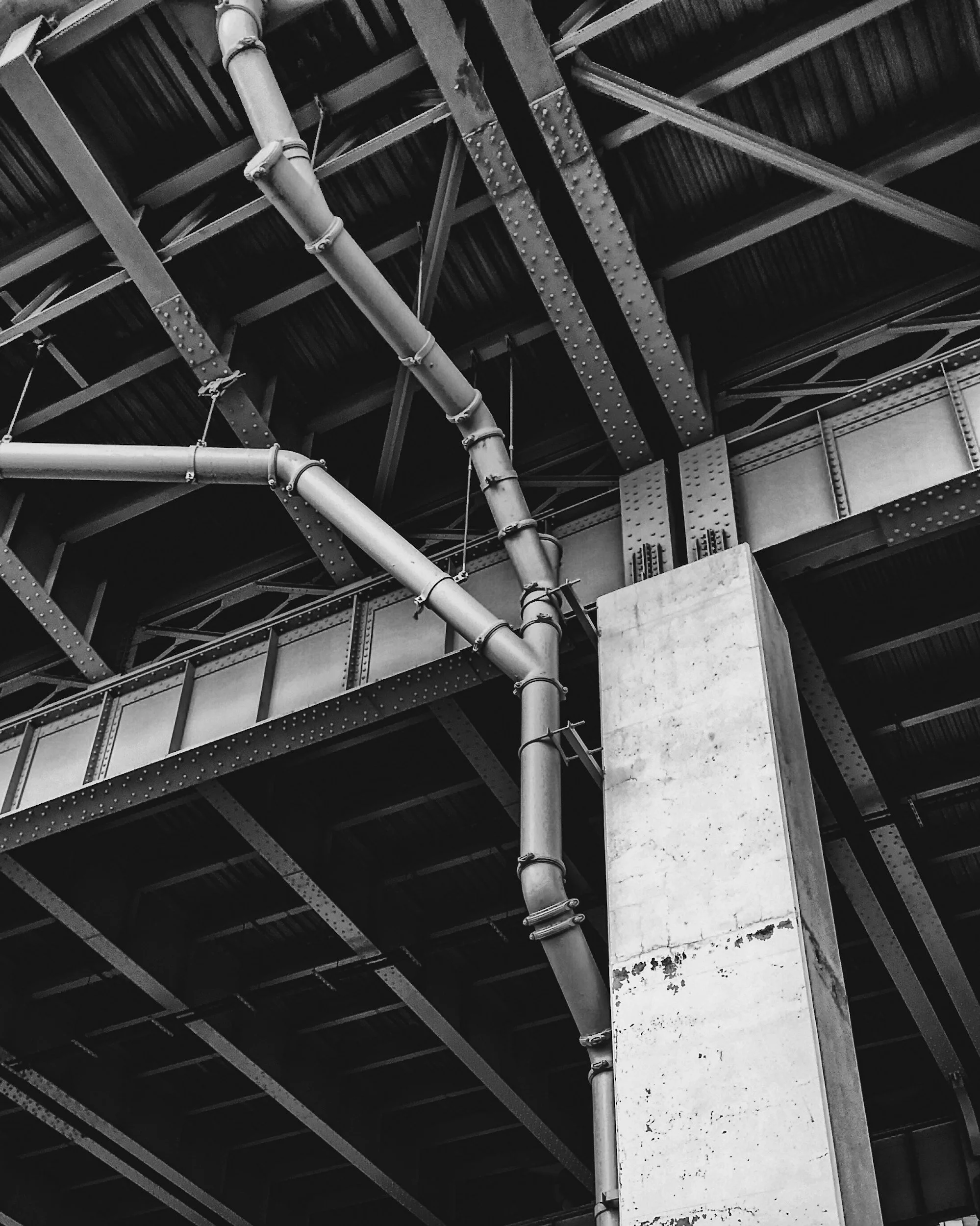 Black and white photo of industrial piping and metal beams under a ceiling, with a concrete pillar supporting the structure.