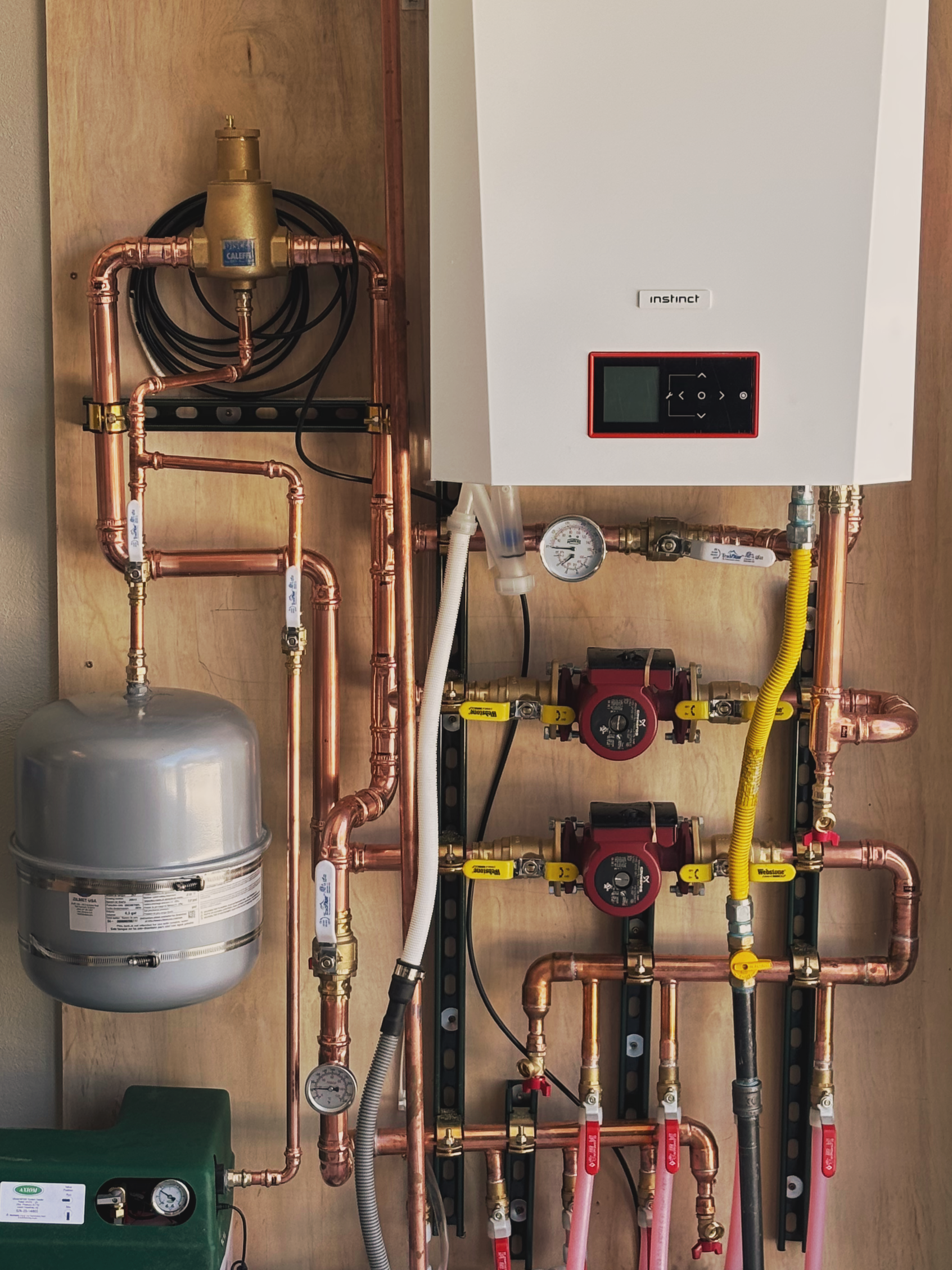 Close-up view of a plumbing and heating system with copper pipes, gauges, a white control box labeled 'Instinct', and various valves, mounted on a wooden panel.