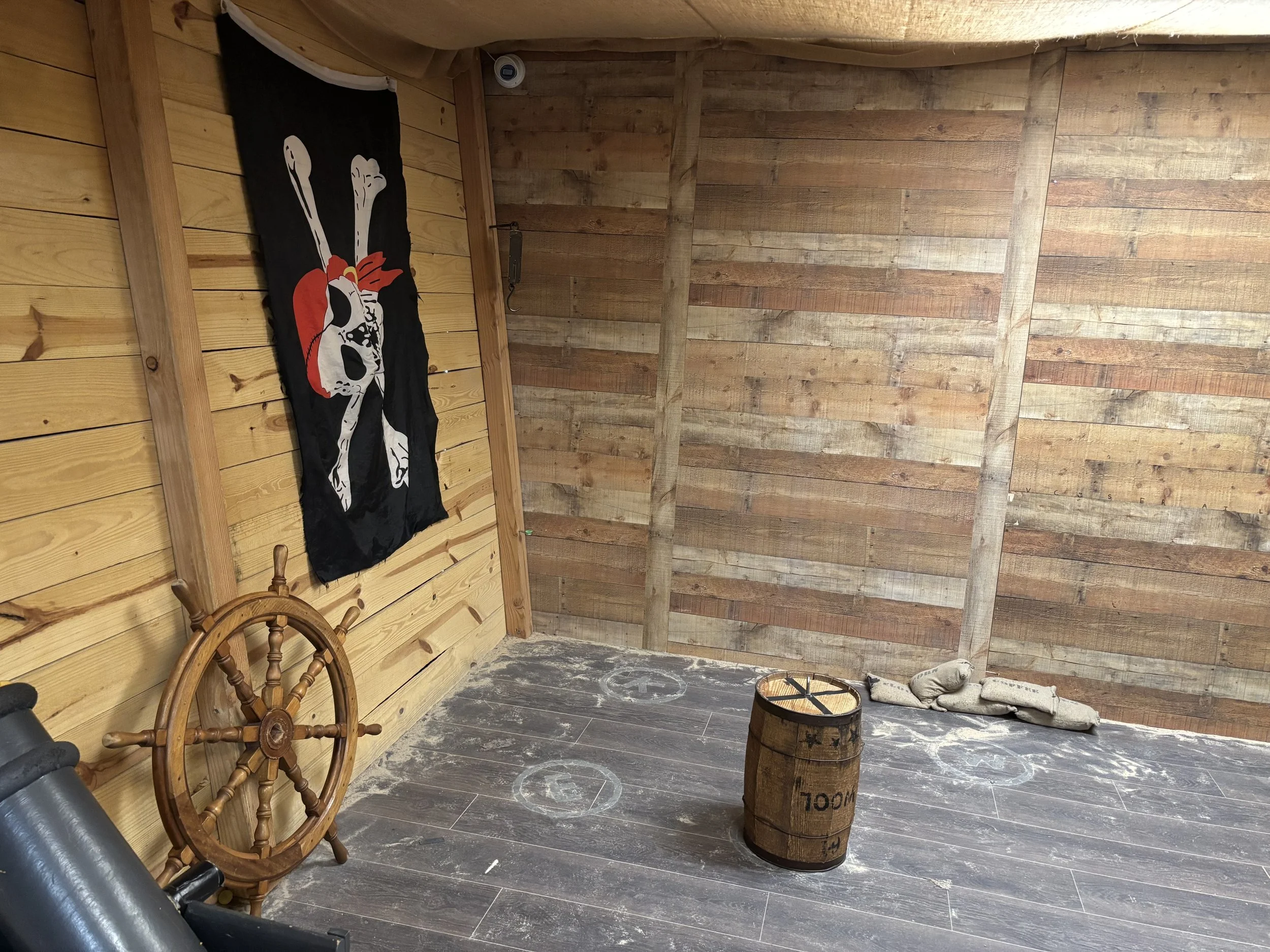 Interior of a wooden room with a pirate flag on the wall, a ship's wheel, a barrel, and a pair of shoes on the floor.