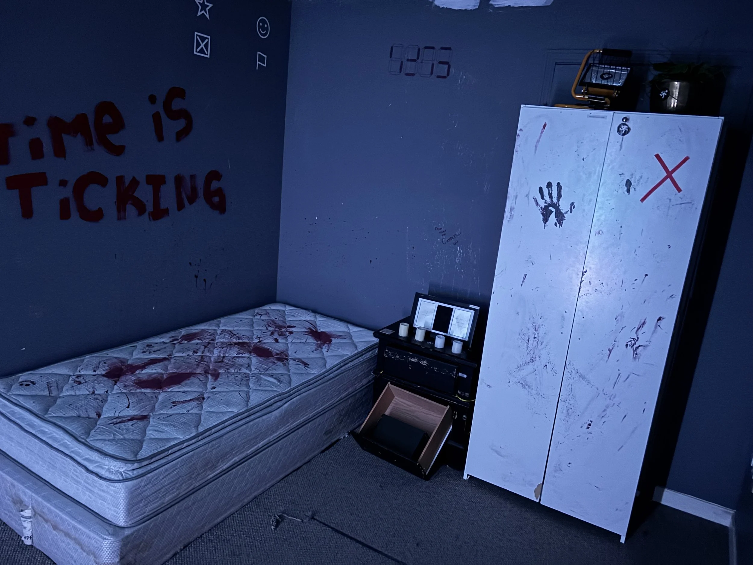 A bedroom with a dark blue wall, containing a mattress with blood stains, a white wardrobe with blood smears and a red 'X' mark, and a small black desk with candles and a picture frame. The wall has red and black graffiti, and the phrase "Time is Ticking" is painted in red. There are handprints and scribbles on the wardrobe and wall.