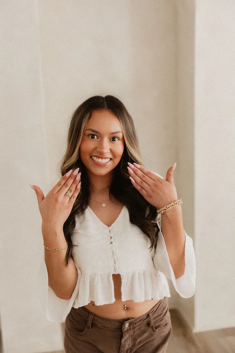 A young woman smiling and showing off her nails, wearing a white crop top, brown pants, jewelry including bracelets, a necklace, and a belly button ring, standing indoors.