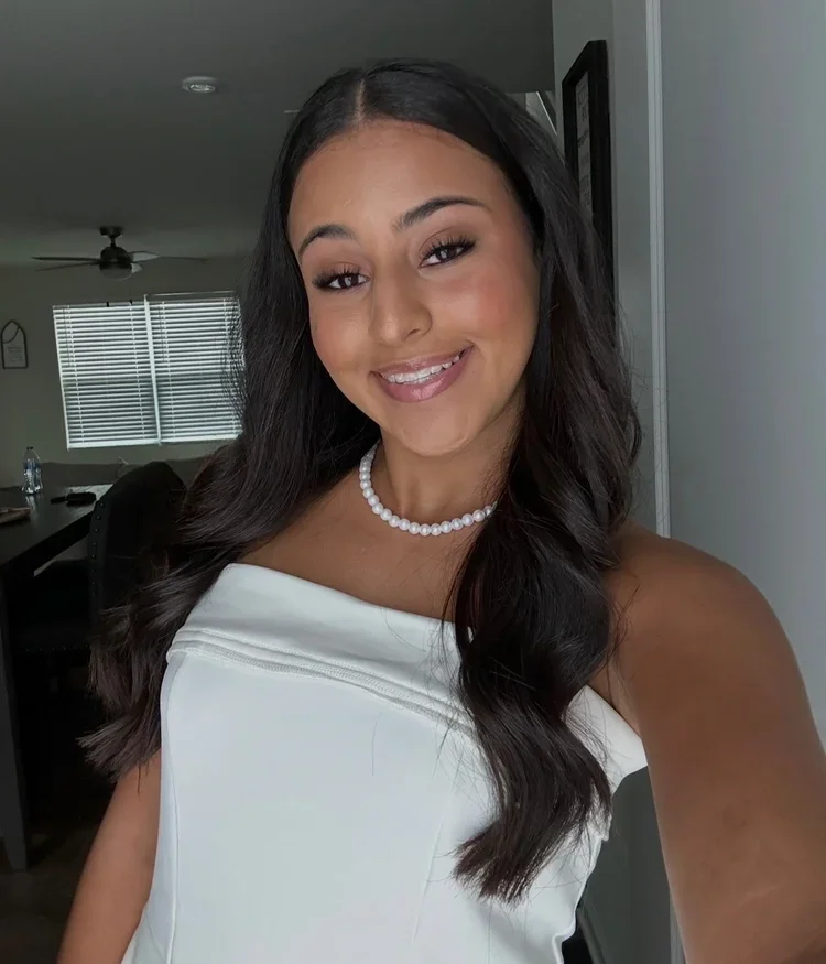 A young woman with long dark hair, smiling, wearing a white strapless dress and a pearl necklace, taking a selfie indoors.
