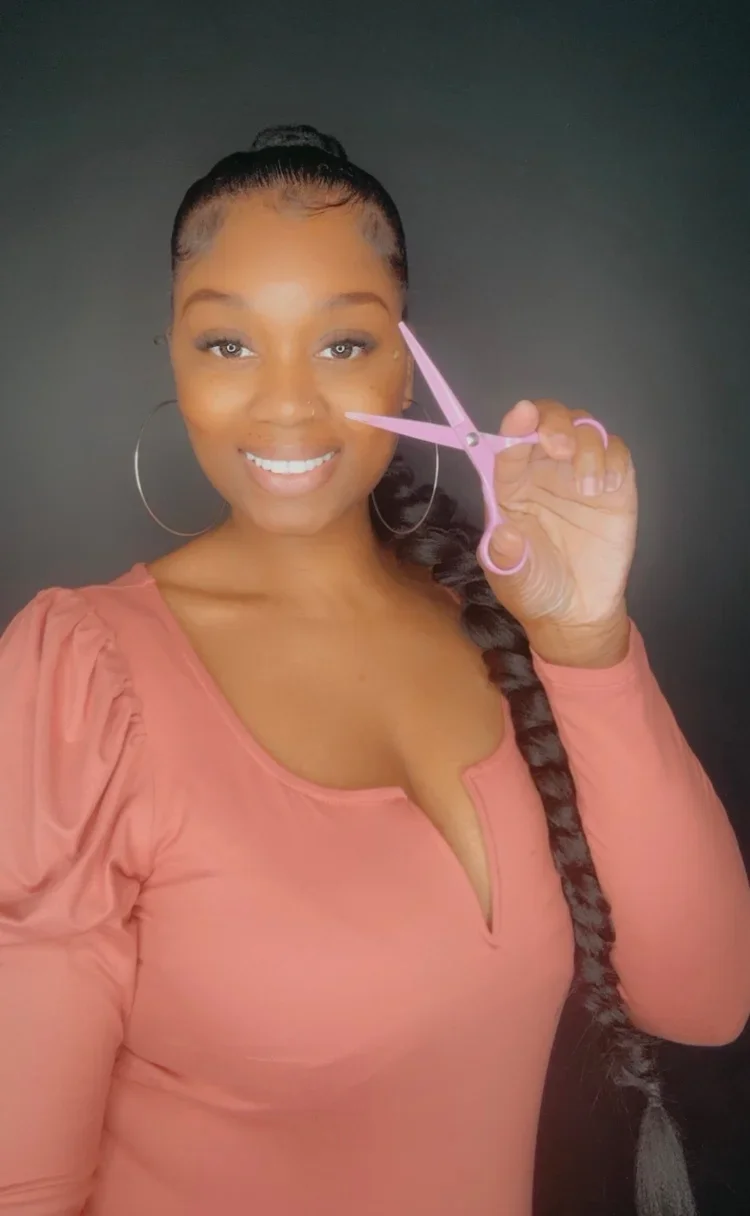 Smiling woman with large hoop earrings holding pink scissors near her face against a black background.