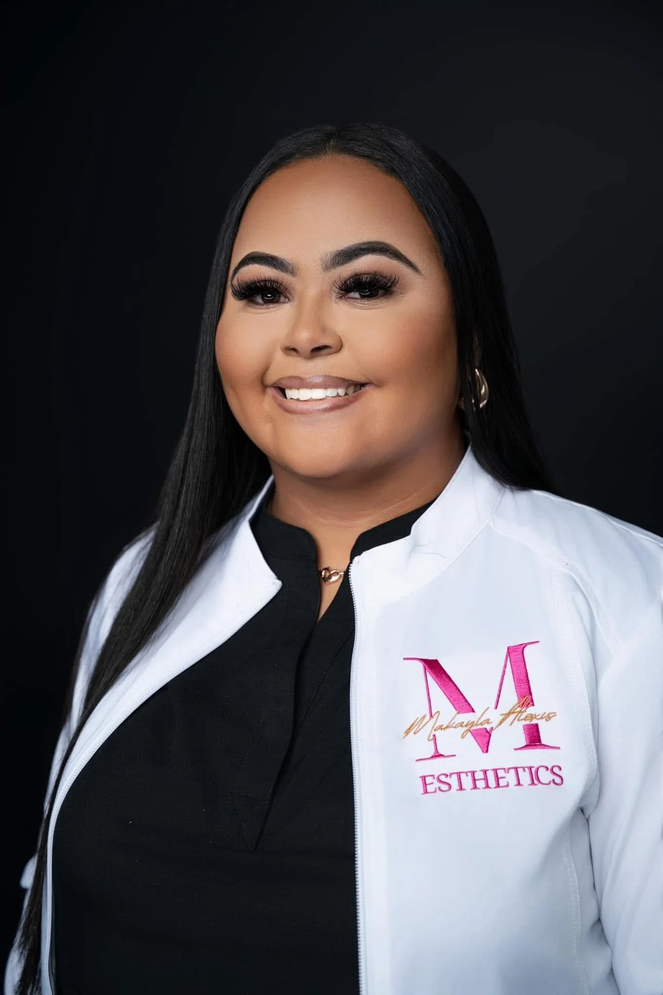 A woman with long dark hair, makeup, and earrings smiling, wearing a white jacket with pink text that reads "Makayla Flowers" and "Esthetics" on it, against a black background.