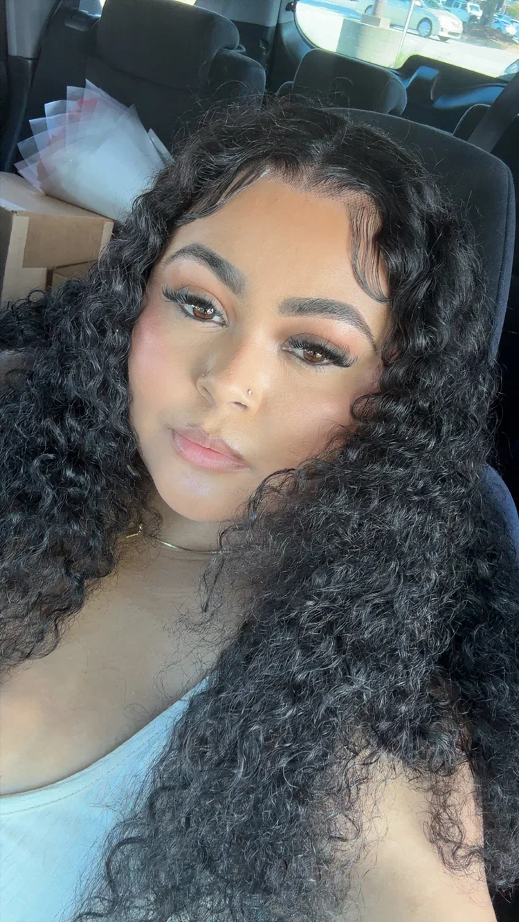 A woman with curly black hair, wearing makeup, with a nose piercing, sitting in a vehicle, with boxes in the background.