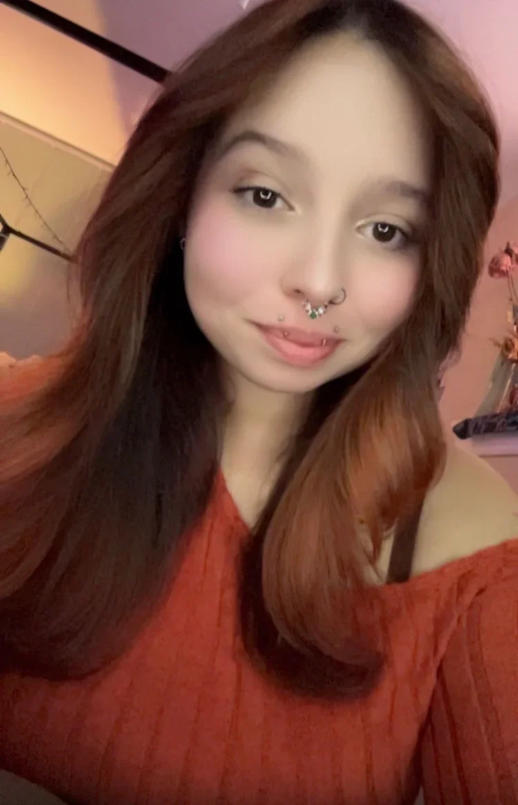 A young woman with long reddish-brown hair taking a selfie in a cozy room. She has a septum piercing, facial piercings, and is wearing an off-the-shoulder red sweater.