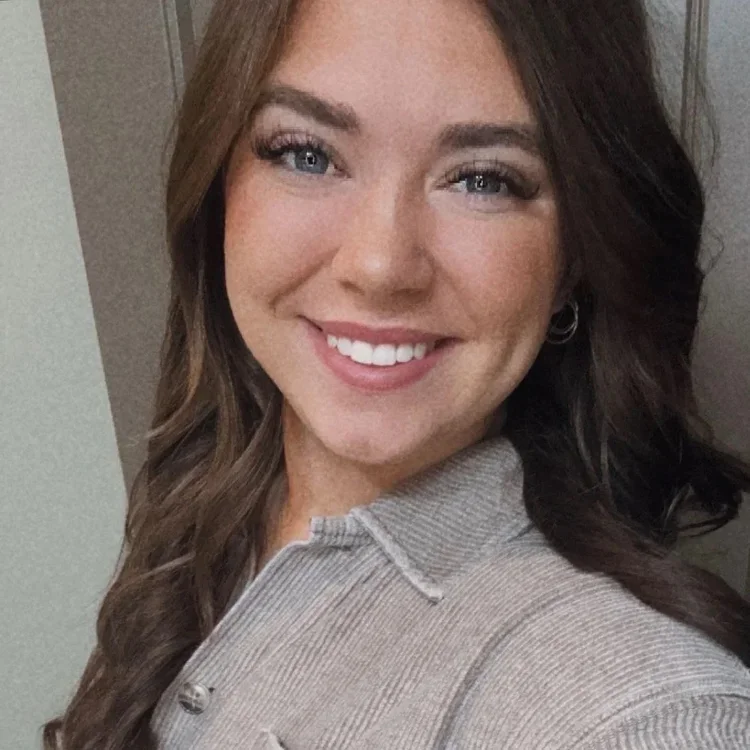 A young woman with long wavy brown hair, blue eyes, and pink lipstick smiling and taking a selfie indoors.