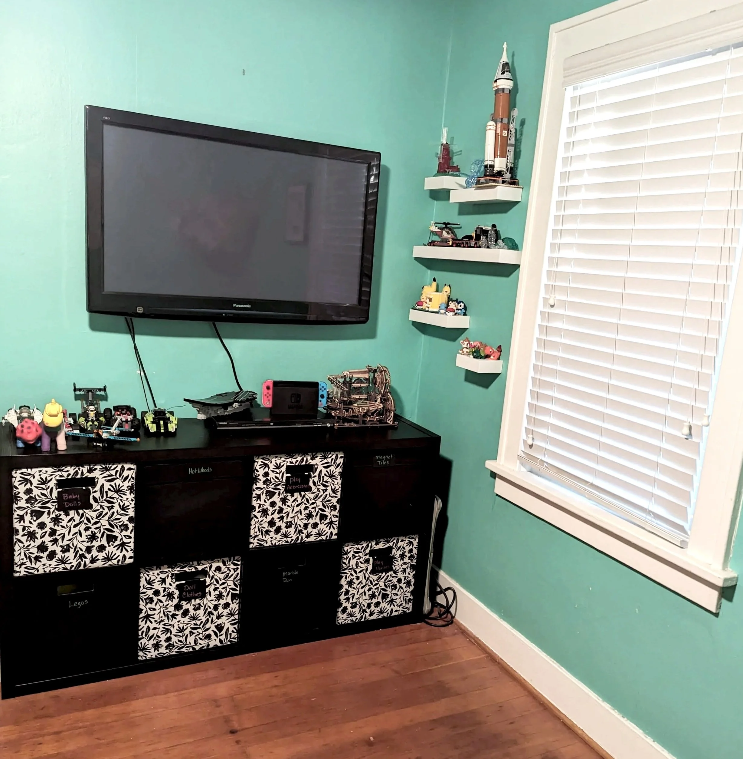 A room corner with a wall-mounted TV, a black cabinet with labeled fabric drawers, and white shelves with model rockets and toys. There are small toy vehicles on the cabinet and a window with white blinds.