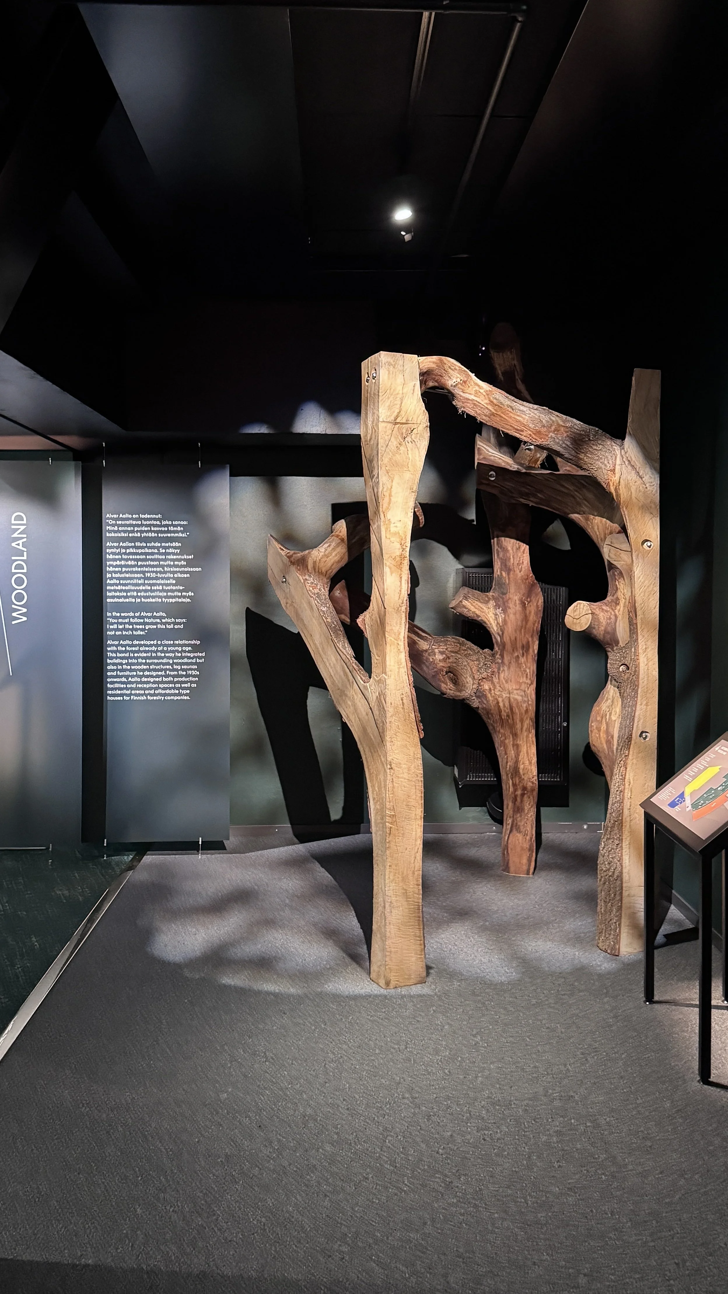 A wooden sculpture displayed in a museum exhibit with informational signage on the left and a small stand with an informational plaque on the right.