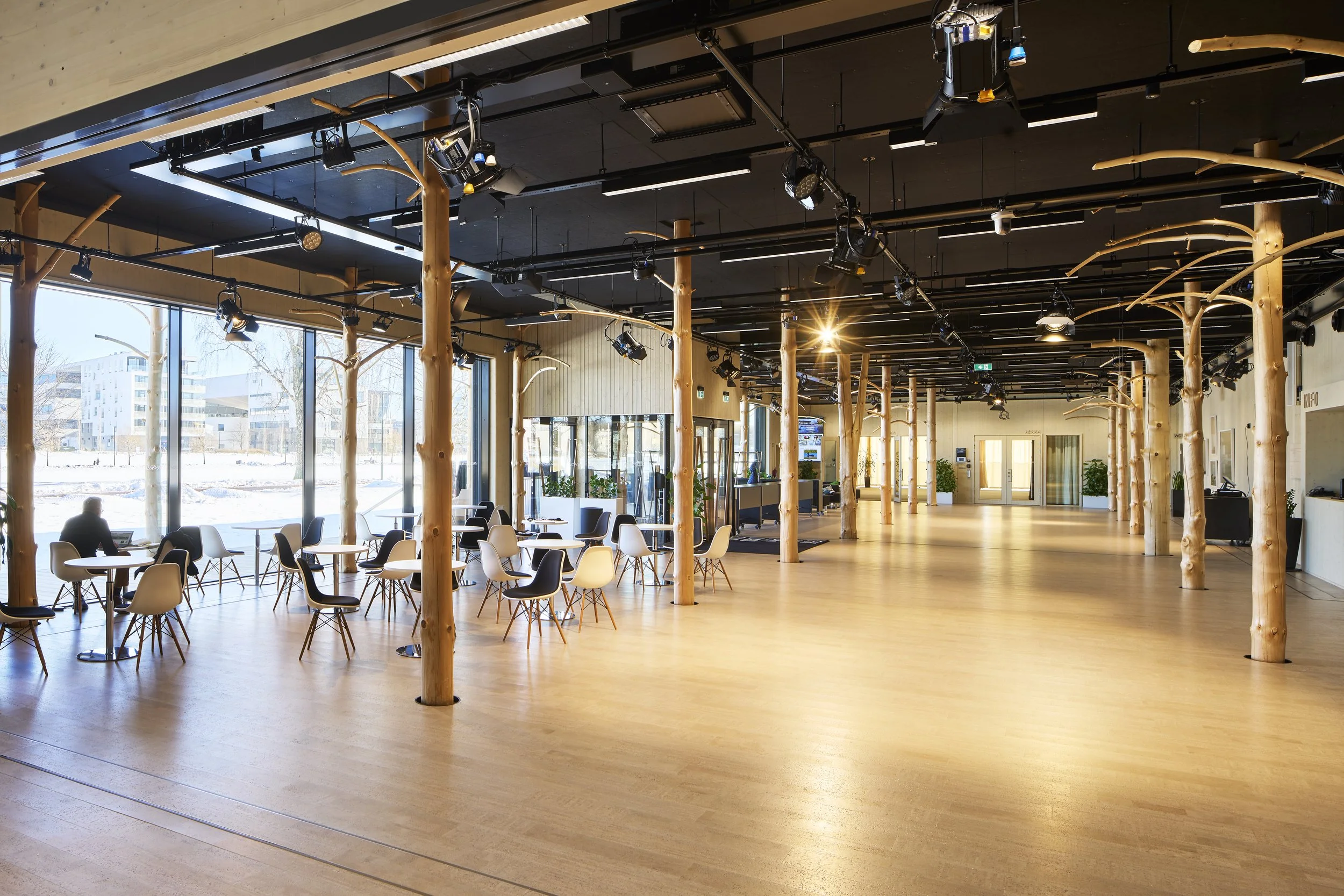 Spacious indoor venue with large windows, wooden floor, numerous white and black chairs and tables, tree-like wooden support columns, modern lighting, with a person sitting by the window and snowy cityscape outside.