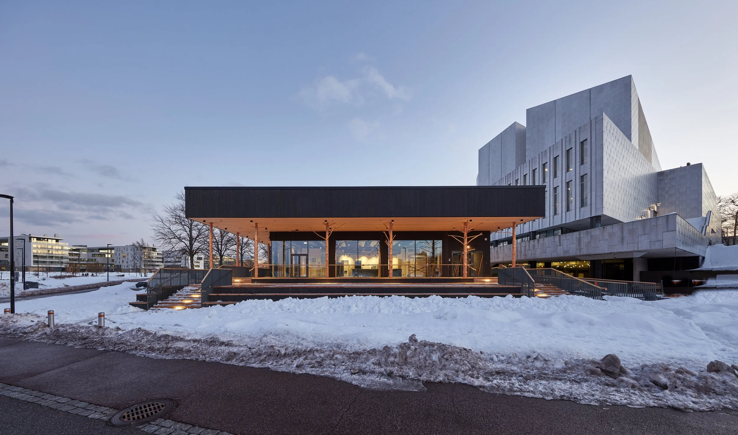 Modern building with an illuminated porch and snow-covered surroundings, clear evening sky.