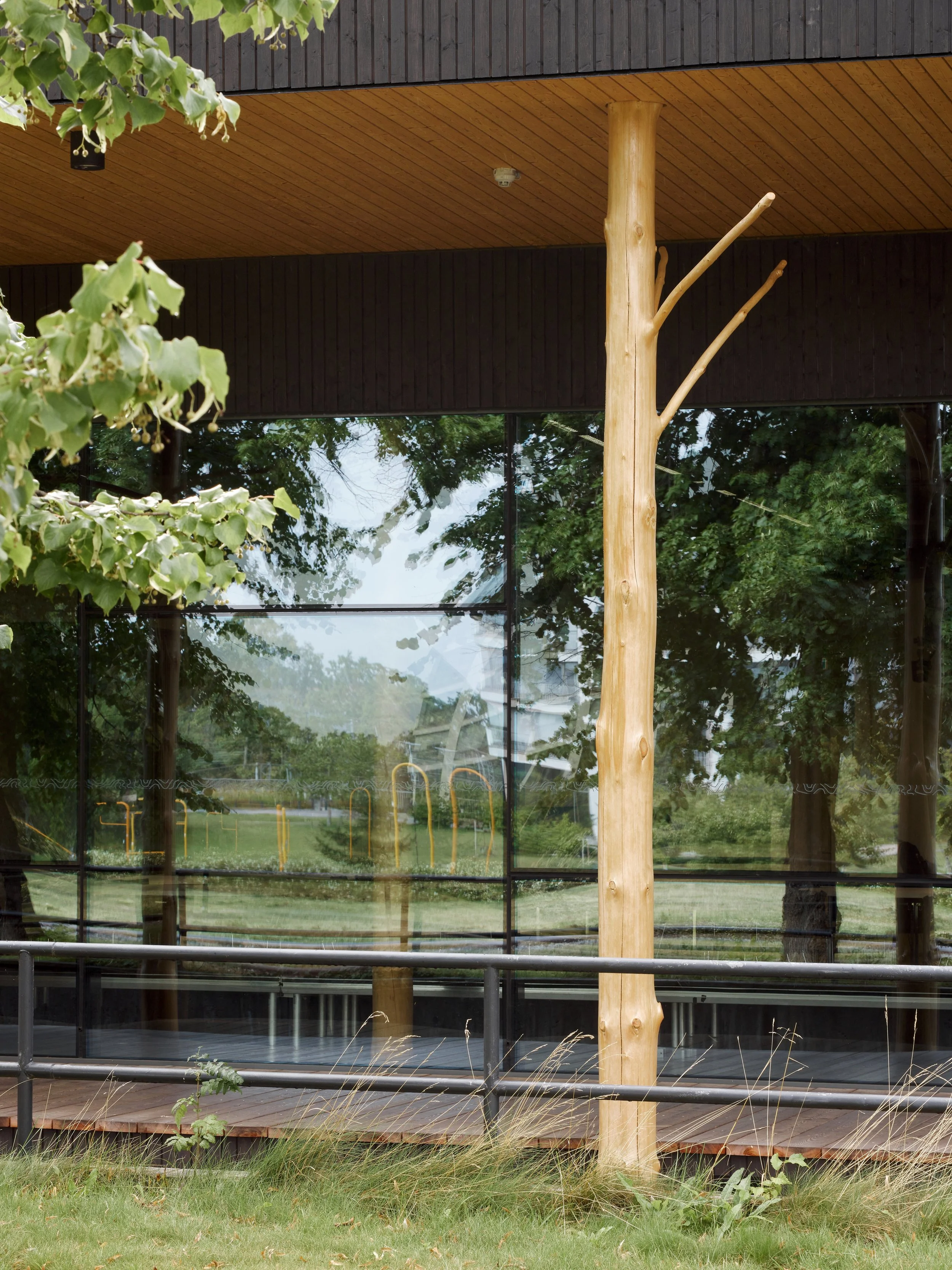 Modern building with large reflective glass windows, wooden deck, and decorative tree-like wooden support pole, surrounded by greenery.