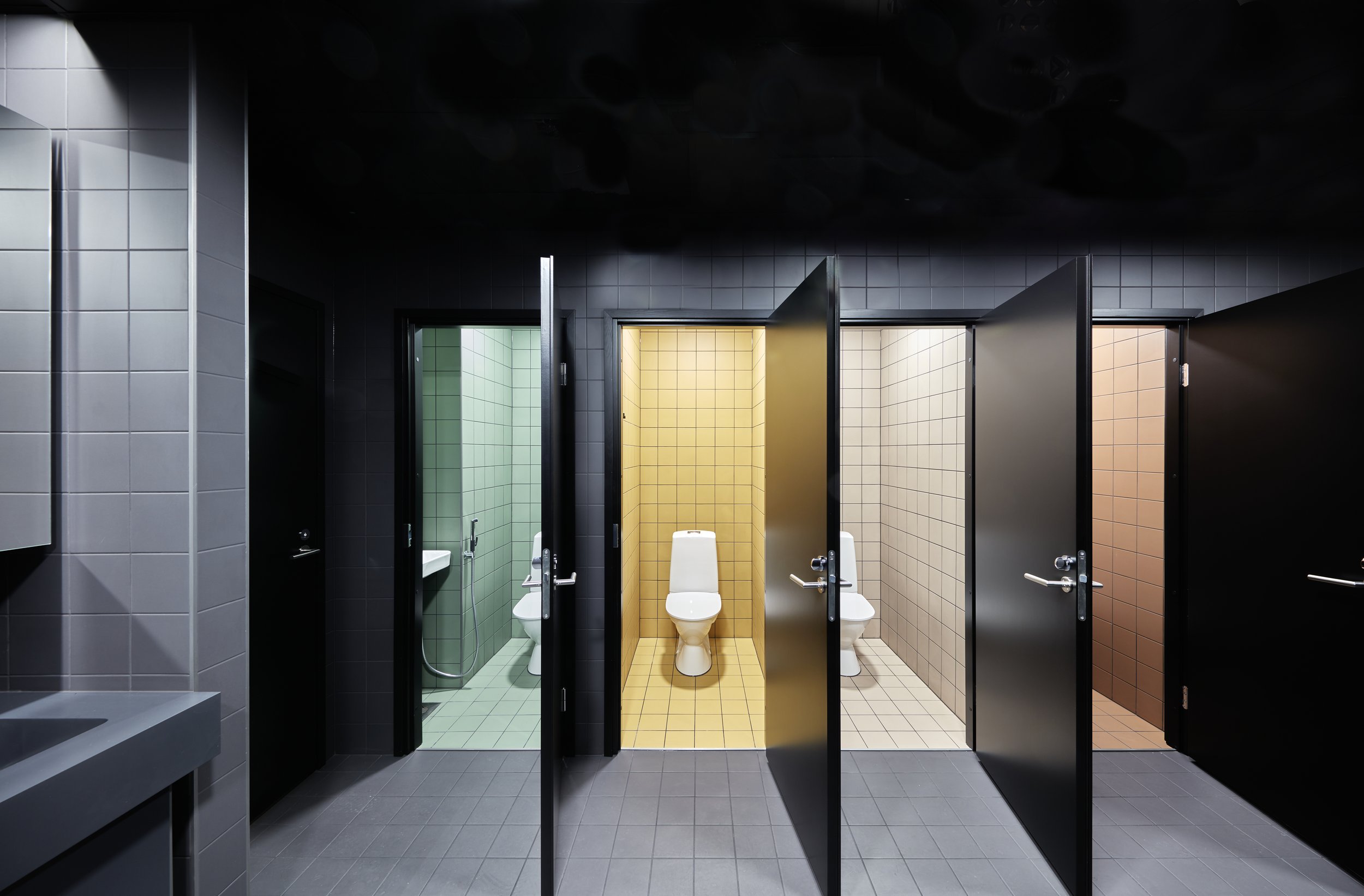 Public restroom with four stalls, each with a different color tile background: green, yellow, beige, and light brown, with black partitions.