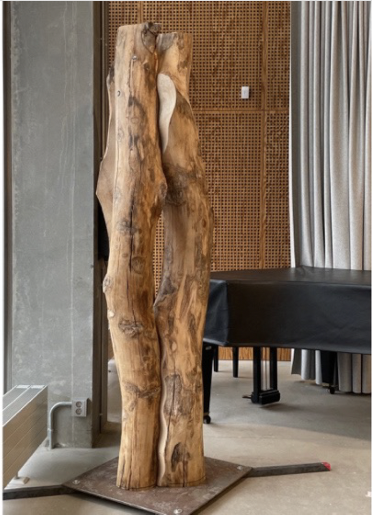 A large, natural wooden sculpture standing indoors next to a table and curtains.