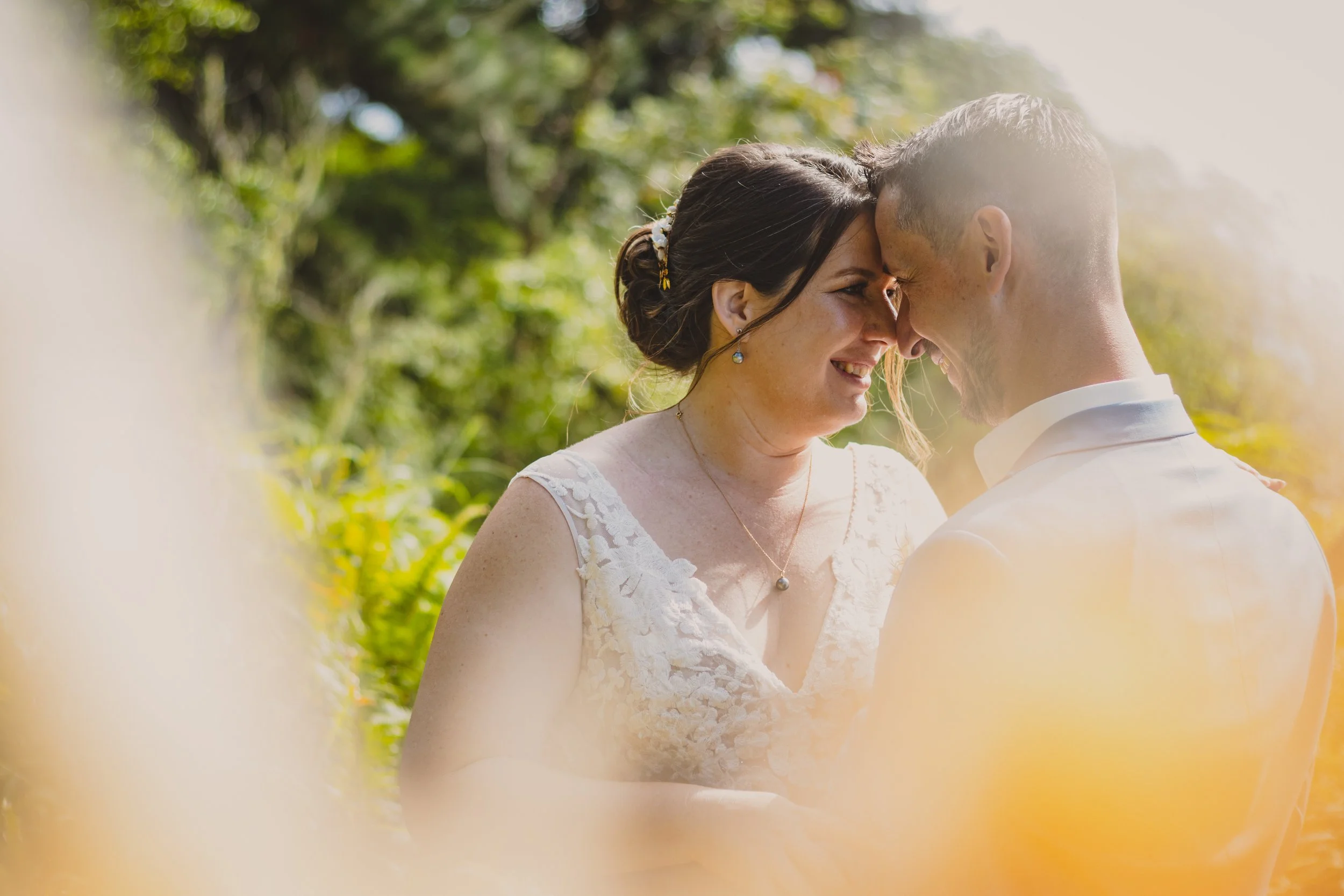 Un couple en mariage, se tenant la tête et se regardant avec des sourires, en plein air avec des arbres en arrière-plan et une lumière douce.