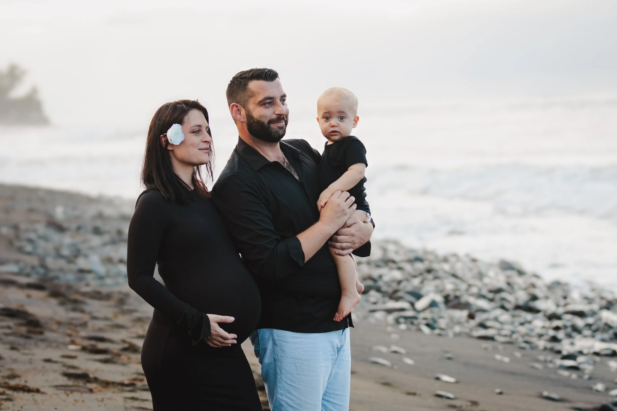 Une famille pose sur la plage, la femme enceinte, l'homme tenant un jeune enfant, tous en vêtements noirs, avec la mer en arrière-plan.