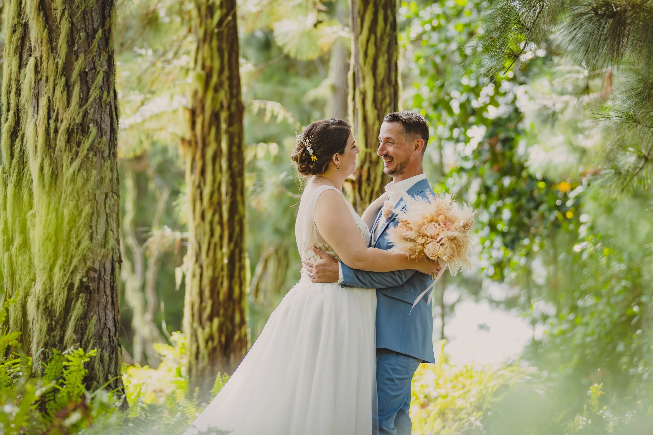 Un couple de mariés danse dans une forêt verdoyante, la femme porte une robe blanche et tient un bouquet de fleurs, l'homme porte un costume bleu, ils se regardent en souriant.