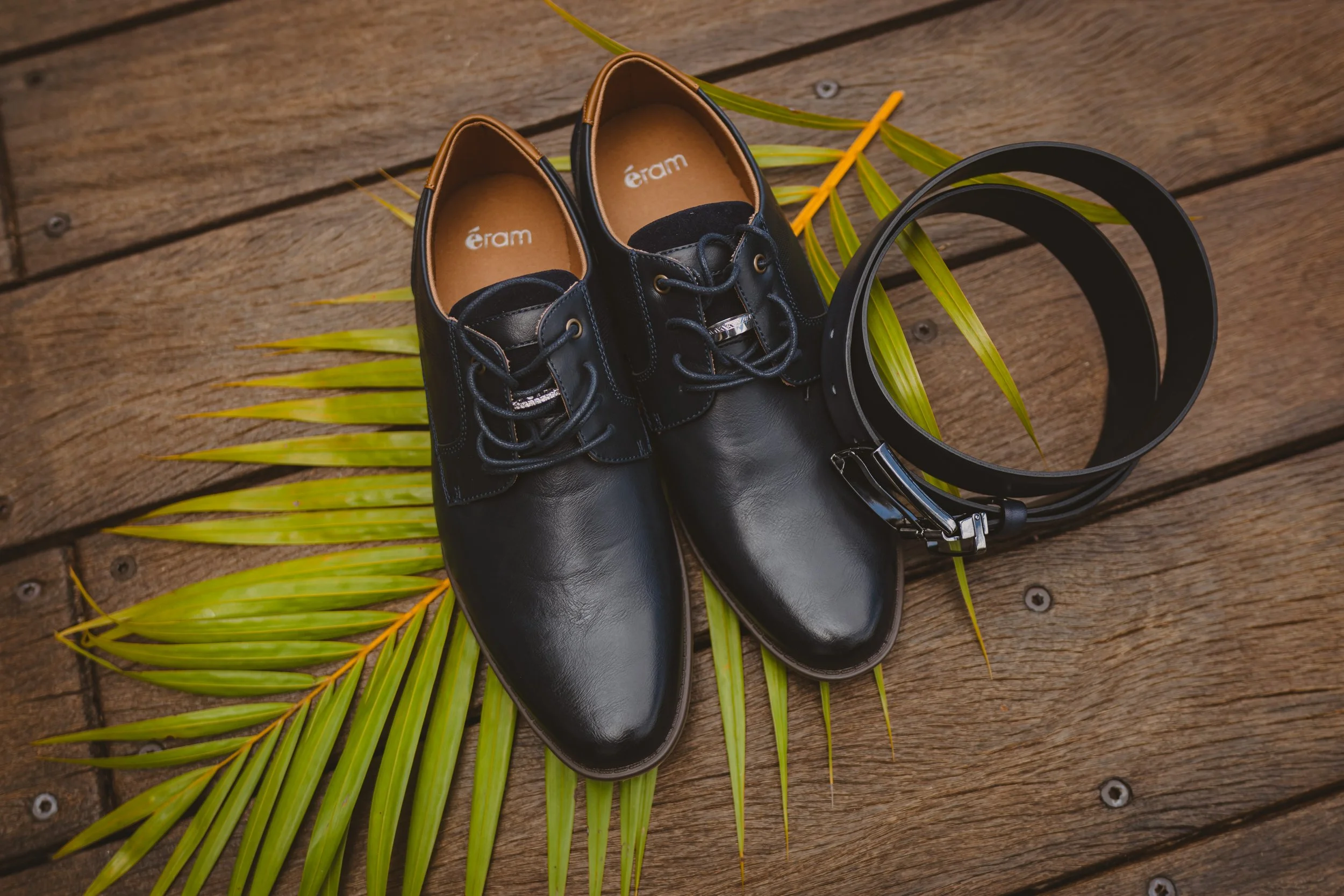 Chaussures en cuir noires, ceinture noire et feuilles vertes sur un sol en bois.