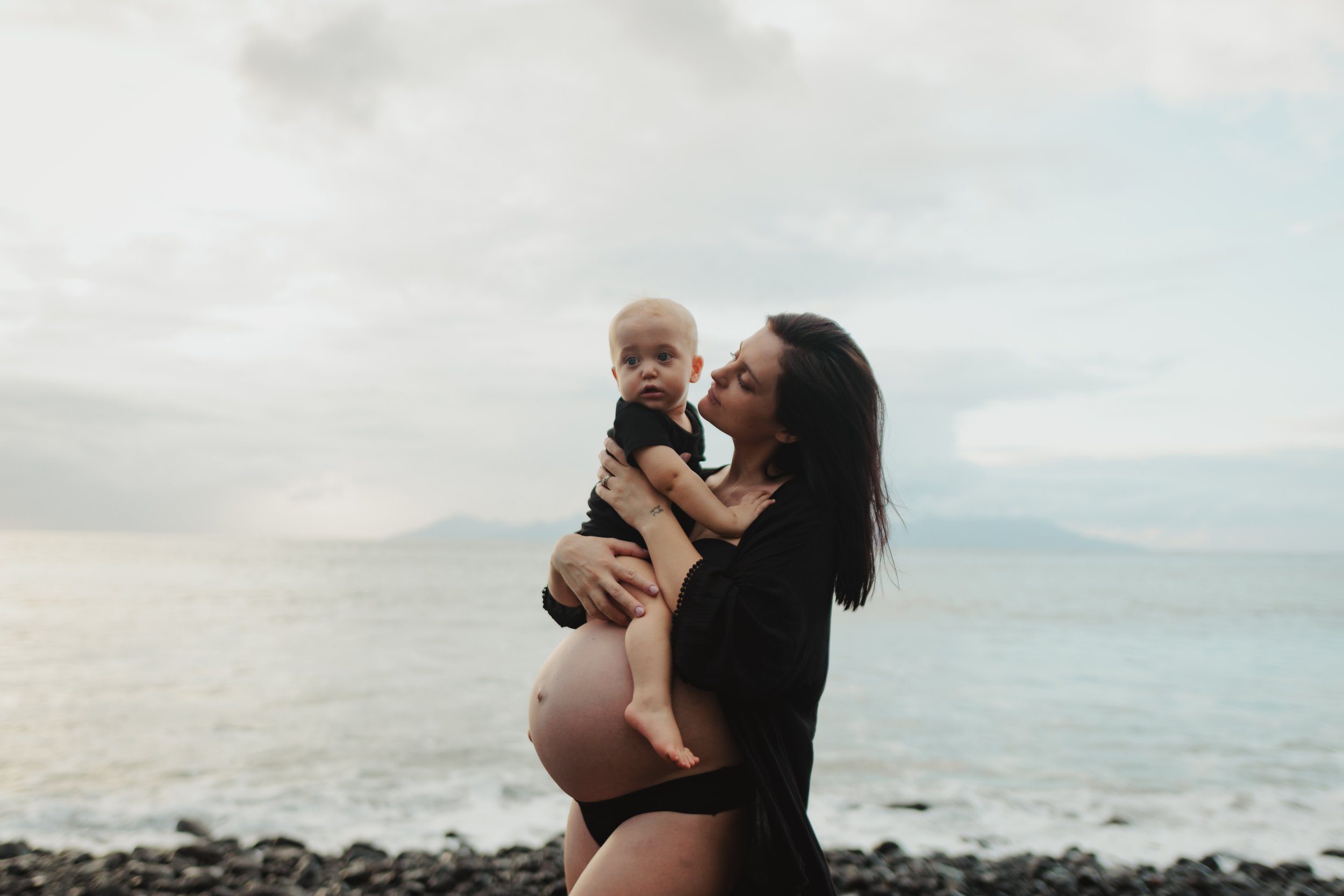 Une femme enceinte tient un bébé dans ses bras sur une plage, vue de profil, avec la mer en arrière-plan, ciel nuageux.